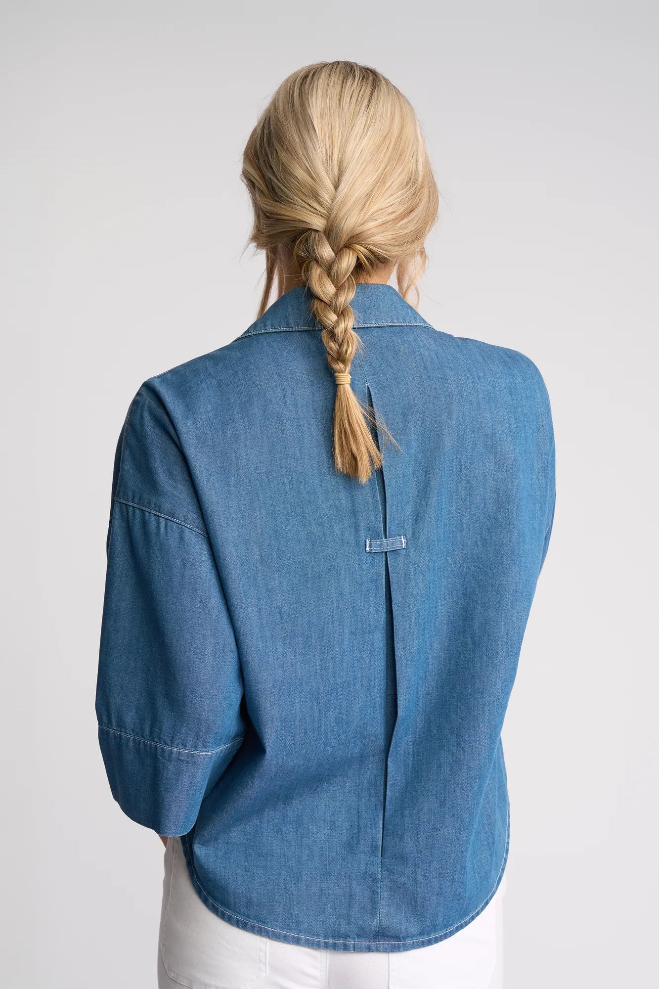 A person with blonde hair in a braid, dressed in the Vassalli Atlas Shirt in Surf Blue and white pants, stands with their back to the camera against a plain light background.