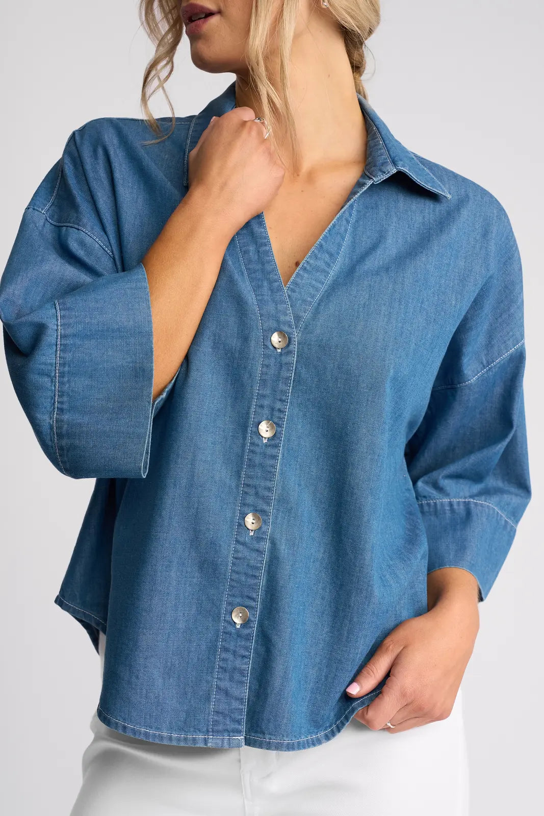 A woman models the Vassalli Atlas Shirt in Surf Blue, a lightweight denim top with wide sleeves, paired with white pants; she stands with one hand touching her neck and the other resting on her hip.