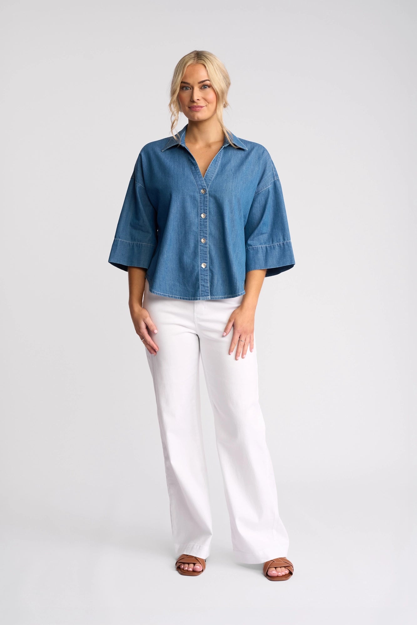 A woman with blonde hair in loose waves stands against a plain background, wearing the Vassalli Atlas Shirt in Surf Blue—a lightweight denim top with three-quarter sleeves—paired with white pants and brown sandals. She has a relaxed expression.