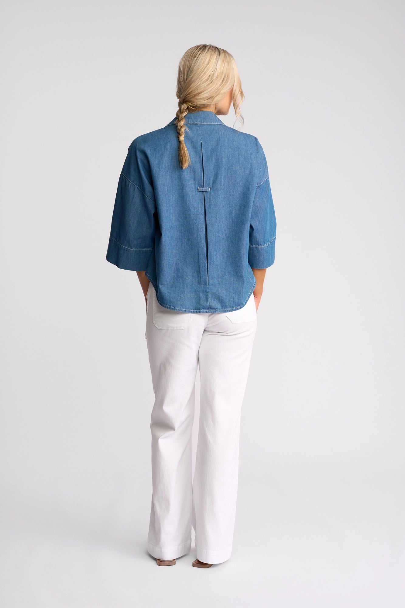 A person with blonde hair in a braid is standing with their back to the camera, wearing the Vassalli Atlas Shirt in Surf Blue and white pants, against a plain light background.