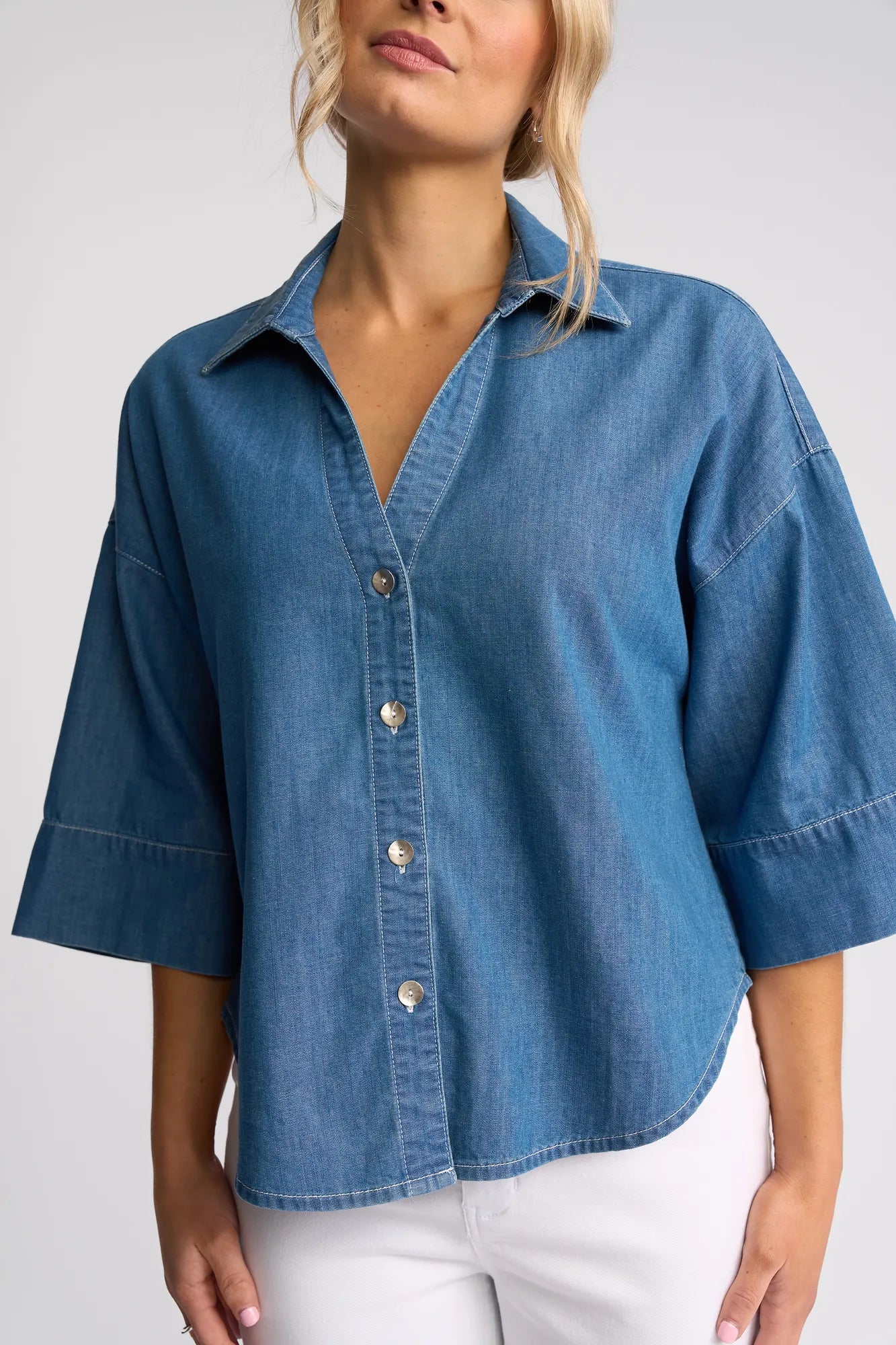 A woman stands against a plain light background, her hair partially tied back, wearing the Vassalli Atlas Shirt in Surf Blue—a loose-fitting, lightweight denim top with wide sleeves—paired with white pants.
