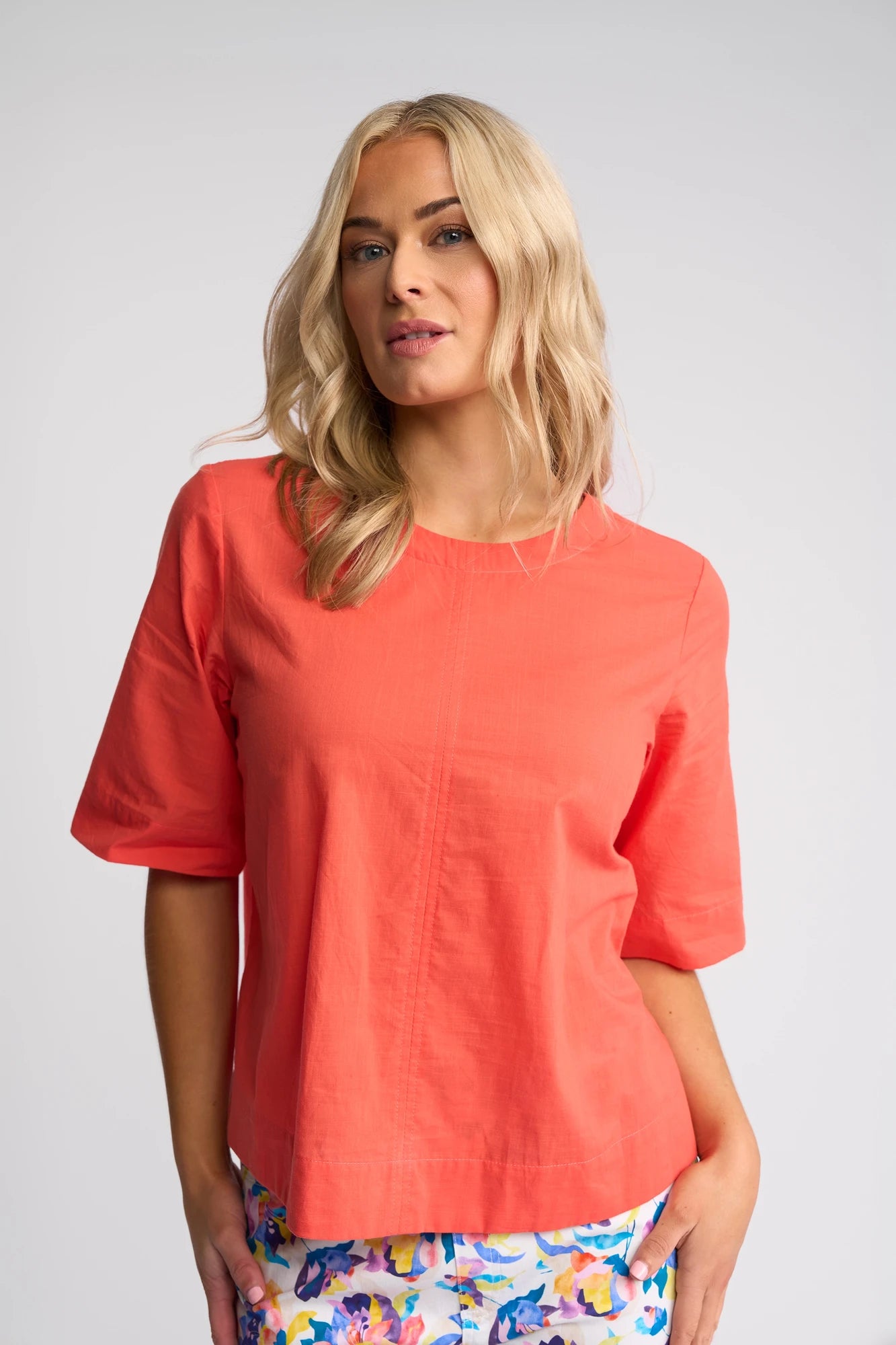 A woman with long blonde hair wears a Vassalli Athena Top in Melon, featuring a boxy fit and 100% cotton fabric, paired with colorful floral pants. She stands against a plain light background and looks slightly to the side.