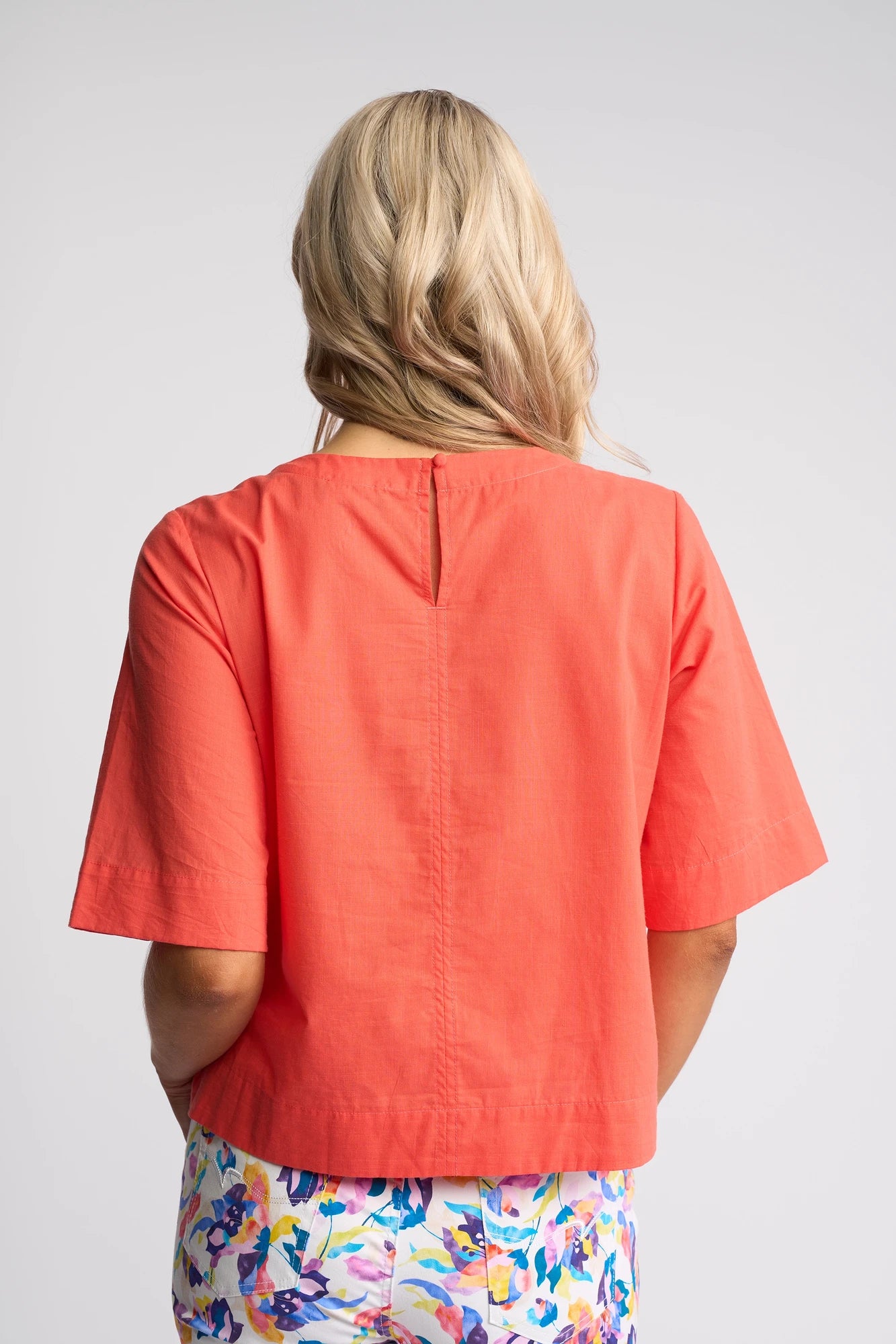 A woman with long blonde hair, shown from the back, wears a short-sleeved Vassalli Athena Top in melon with a boxy fit and colorful floral pants against a light gray background.