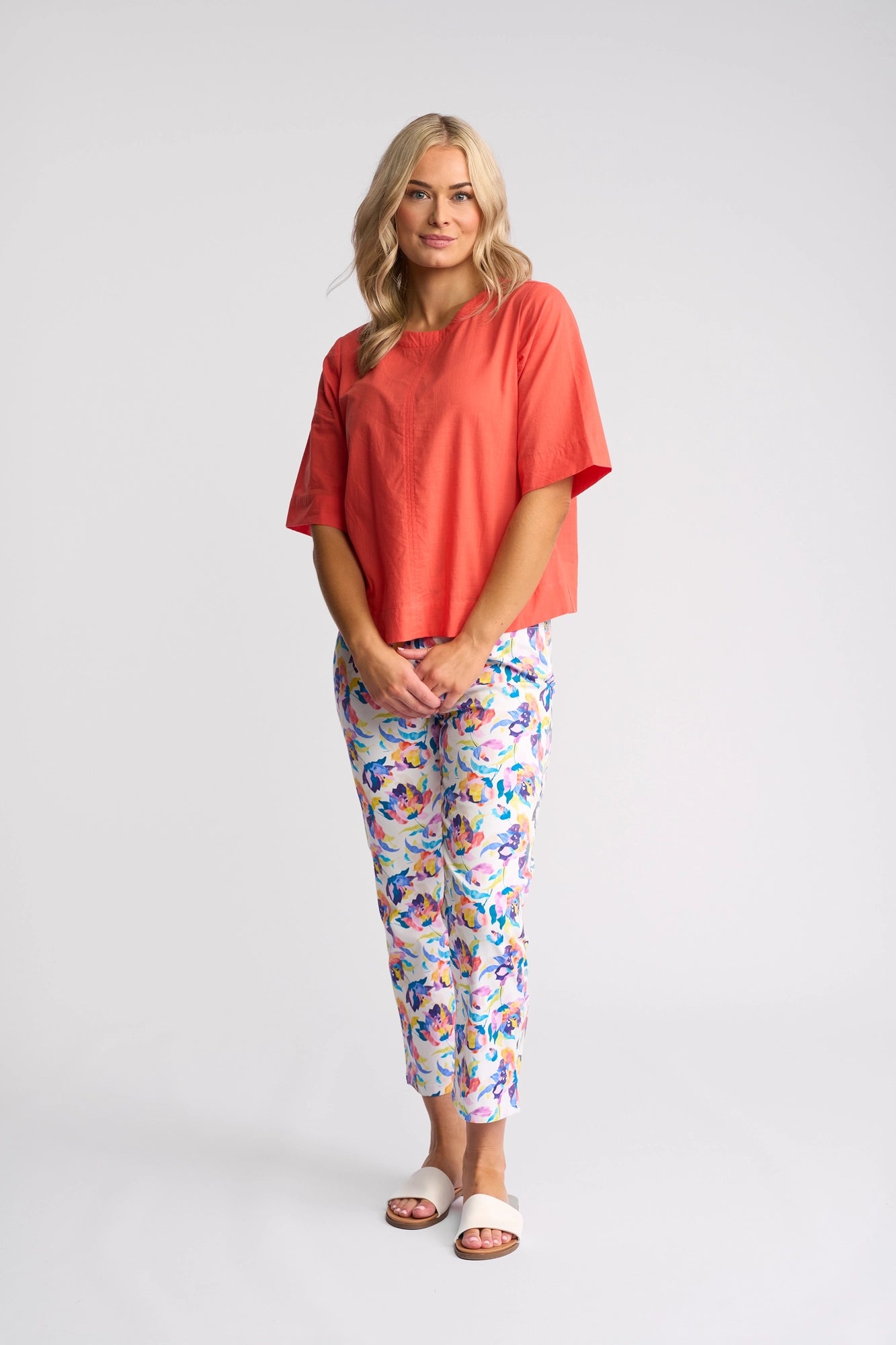A woman stands in a studio wearing the Vassalli Athena Top in Melon, which showcases a loose, boxy silhouette and is made from 100% cotton. She pairs it with vibrant floral patterned pants and white slide sandals, her hands together as she smiles gently.