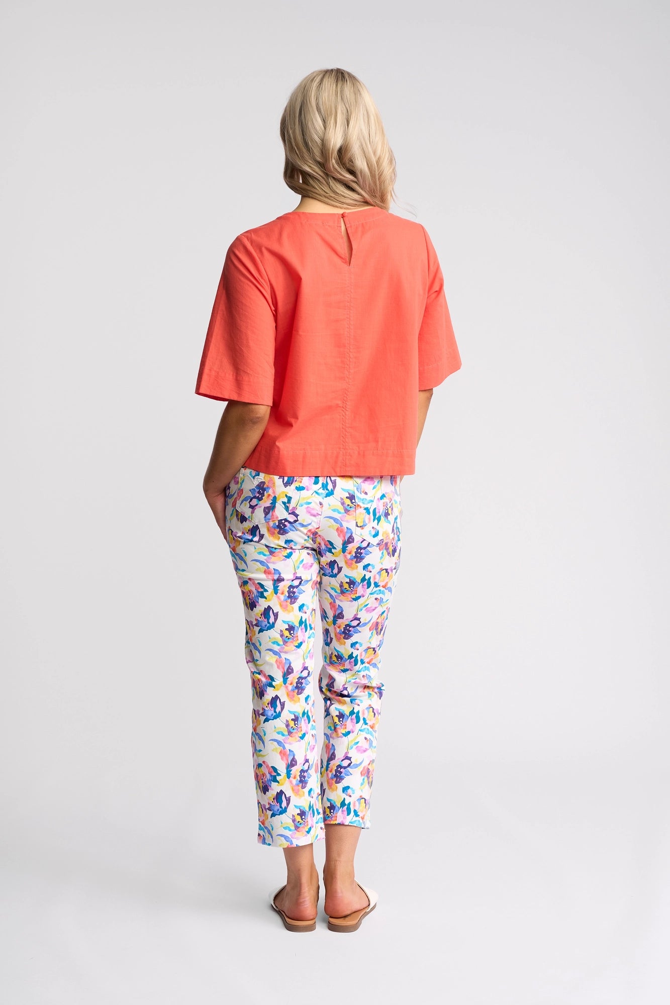 A person with blonde hair stands facing away, wearing a short-sleeve Vassalli Athena Top in Melon with a boxy fit, paired with colorful floral pants and sandals against a plain white background.