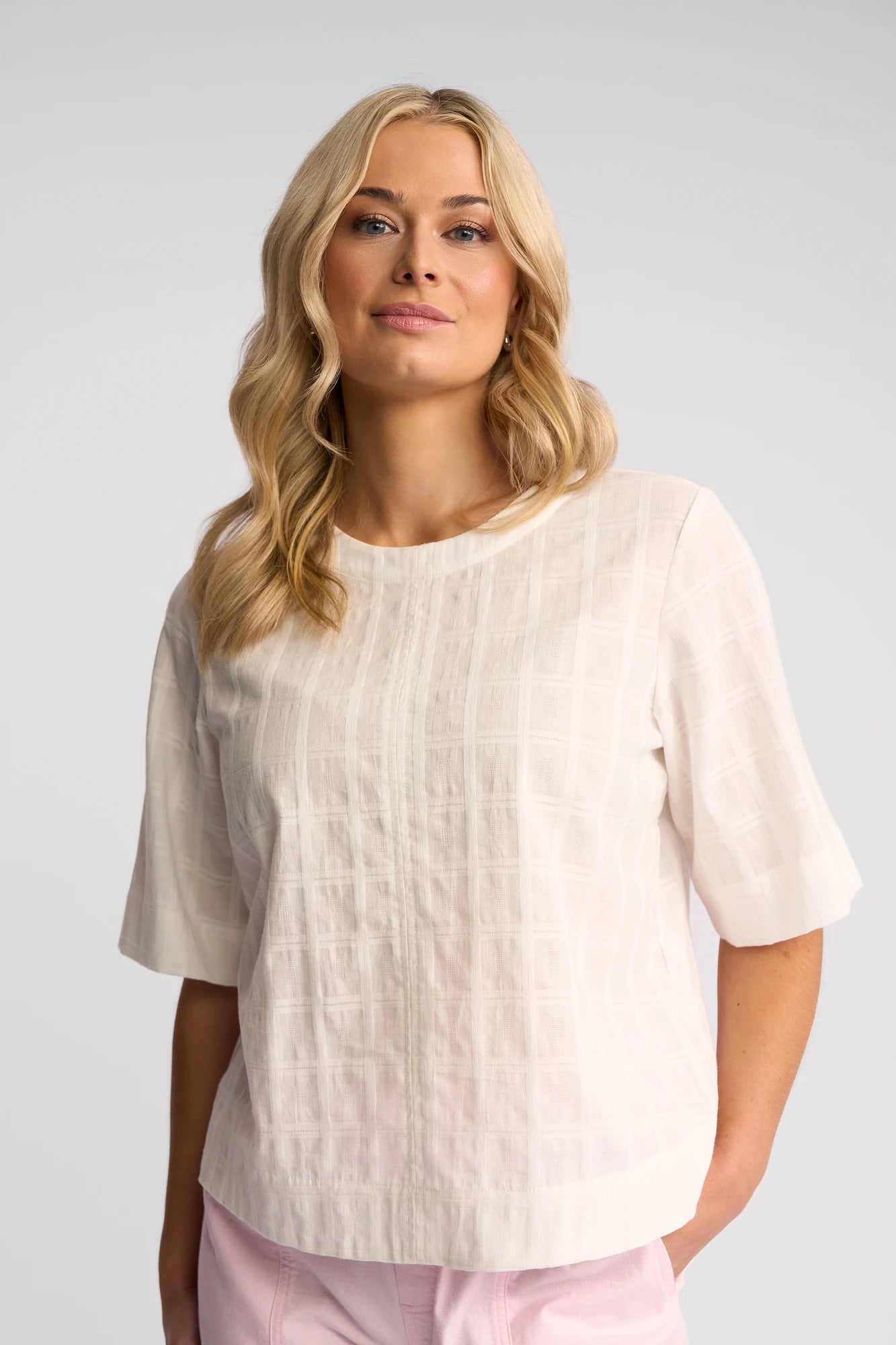 A woman wearing the Vassalli Athena Top - White Check—a 100% cotton textured blouse with short sleeves—paired with light pink pants stands against a plain light background, looking at the camera and smiling softly.