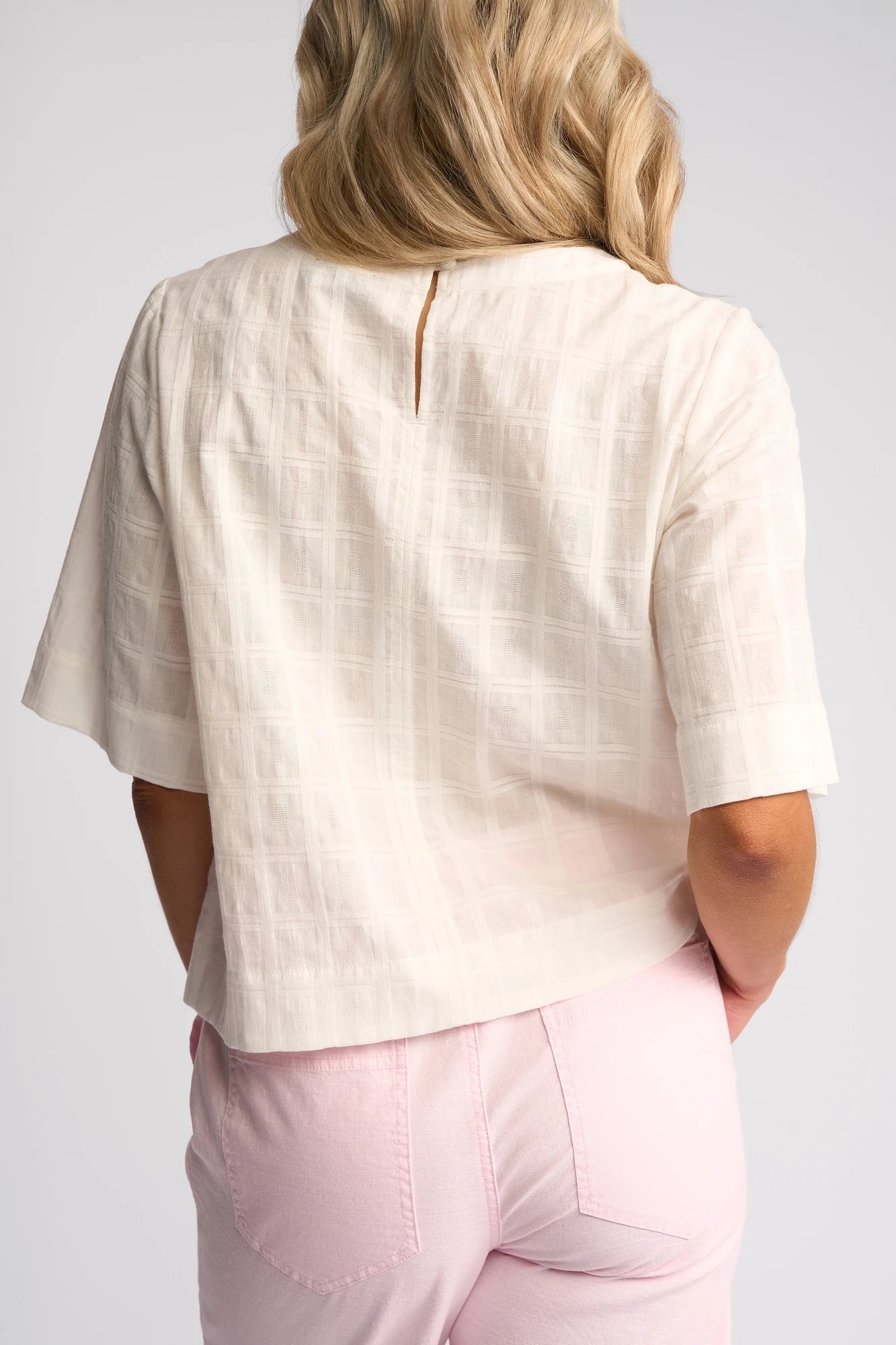 A person with wavy blonde hair is seen from the back against a plain light background, wearing the Vassalli Athena Top in White Check—a short-sleeve blouse made from 100% cotton—and light pink pants.