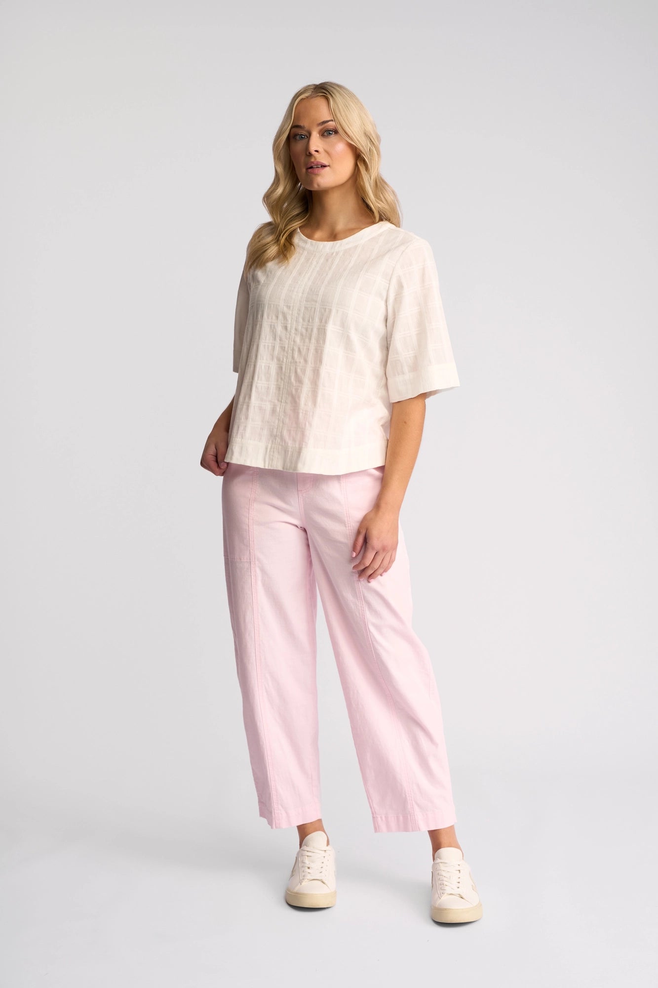 A woman stands against a plain background, wearing the Vassalli Athena Top - White Check crafted from 100% cotton. She pairs it with light pink wide-leg pants and white sneakers. Her blonde hair is loose, and she strikes a relaxed pose with one hand in her pocket.