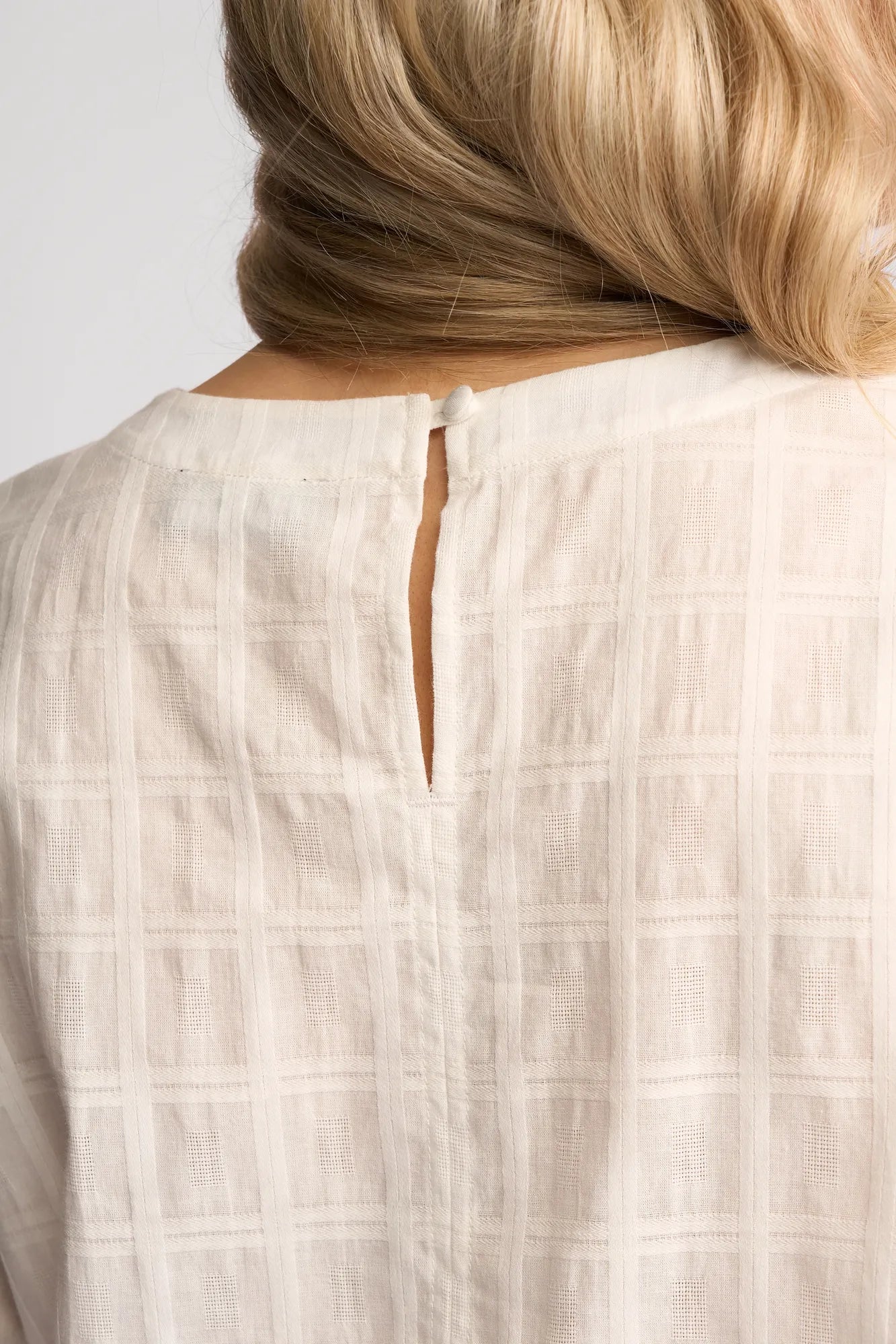 Close-up of the back of the Vassalli Athena Top in White Check, a light-colored, textured blouse made from 100% cotton. The subtle check pattern is complemented by a small slit and button closure at the neckline, while blonde hair partially covers the upper right corner, drawing attention to the garment’s refined details.