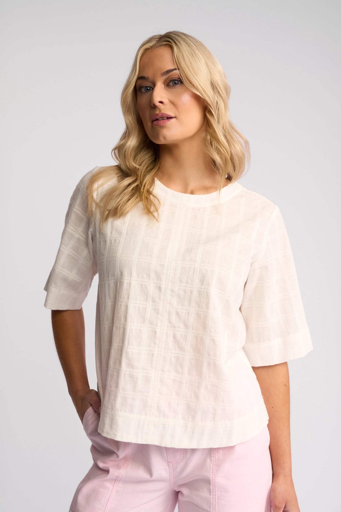 A woman with long blonde hair models the Vassalli Athena Top - White Check, a textured short-sleeve top crafted from 100% cotton; she stands against a plain light background with one hand in her pocket, pairing the piece with light pink pants.