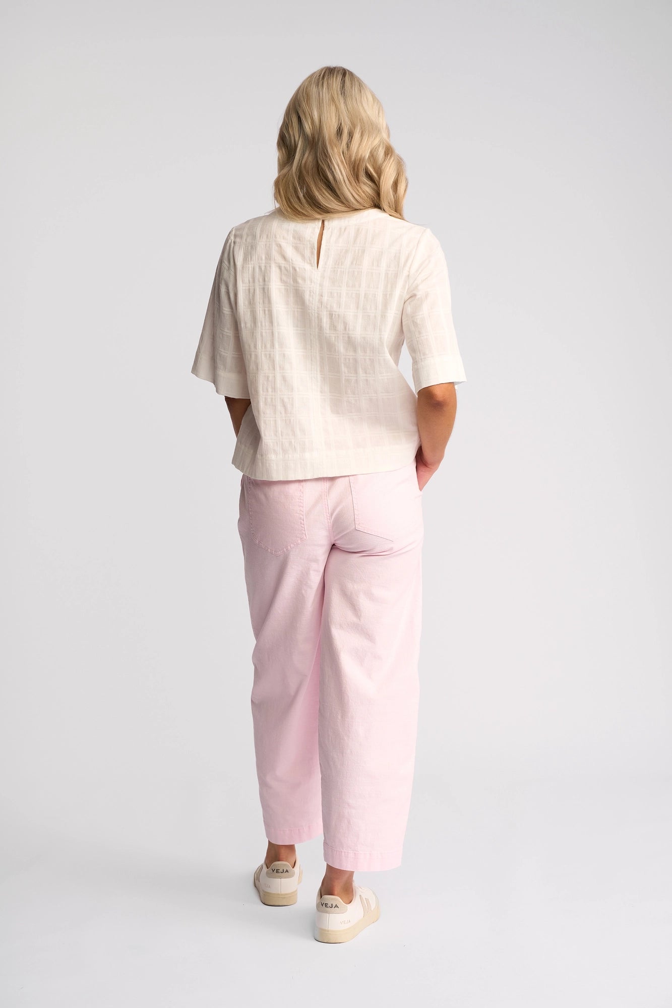 A woman with blonde hair stands facing away against a plain white background, wearing the Vassalli Athena Top in White Check, crafted from 100% cotton. She pairs it with light pink wide-leg pants and beige shoes.