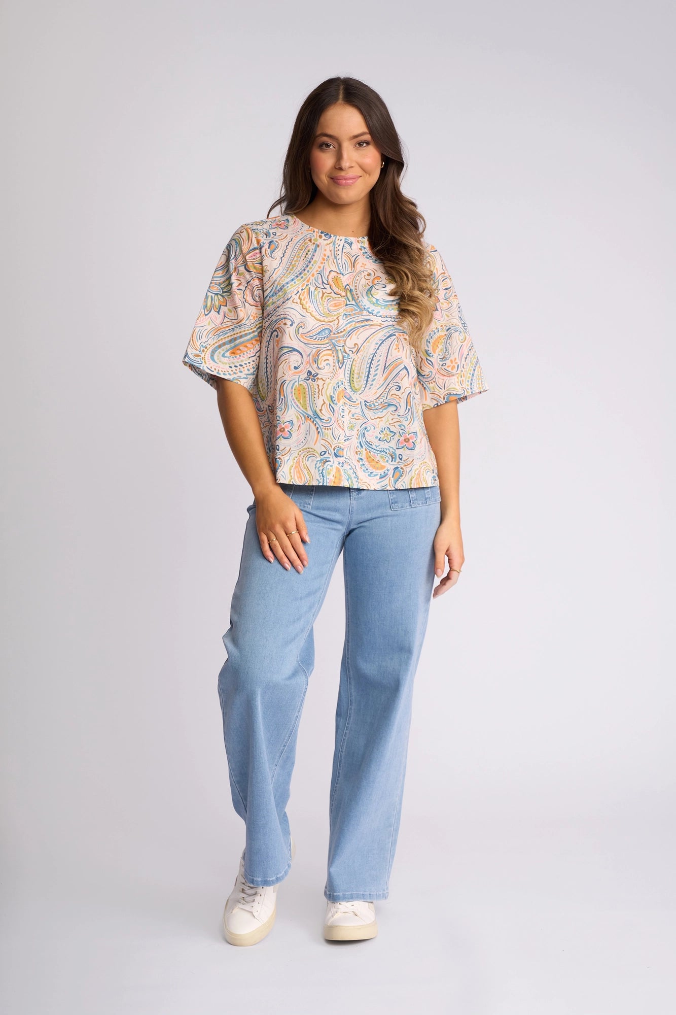 A woman stands against a plain background, wearing the Vassalli Athena Top - Aurora—a boxy cotton top featuring a vibrant aurora print—paired with light blue jeans and white sneakers. She has long, wavy hair and a slight smile.