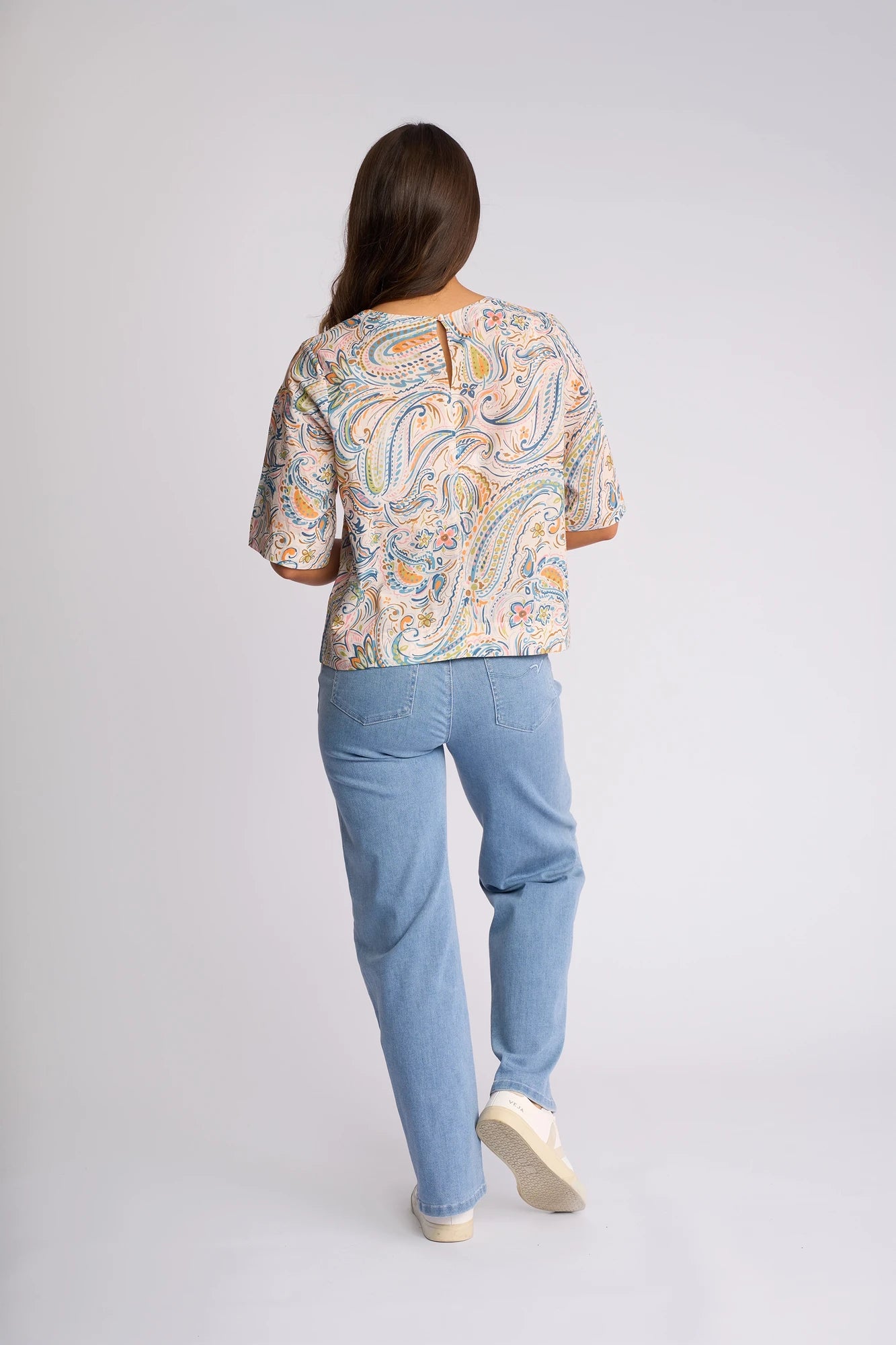 A person with long brown hair, wearing the Vassalli Athena Top - Aurora—a patterned, short-sleeve cotton boxy top—along with light blue jeans and white shoes, stands facing away from the camera against a plain background.