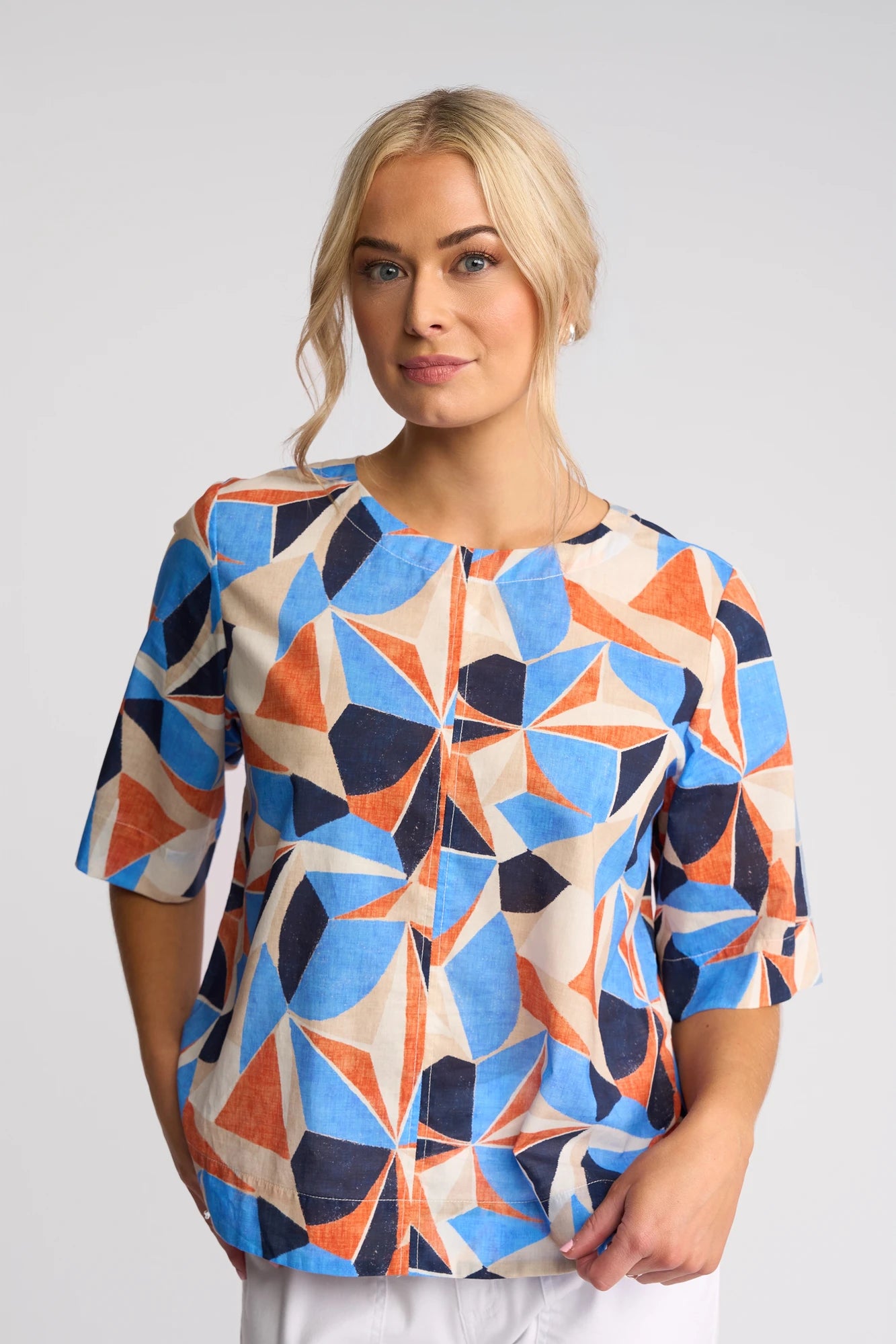 A woman with blonde hair wears the Vassalli Athena Top - Row, featuring a blue, orange, and beige geometric print in cotton and a boxy fit, standing against a light gray background with a neutral expression.