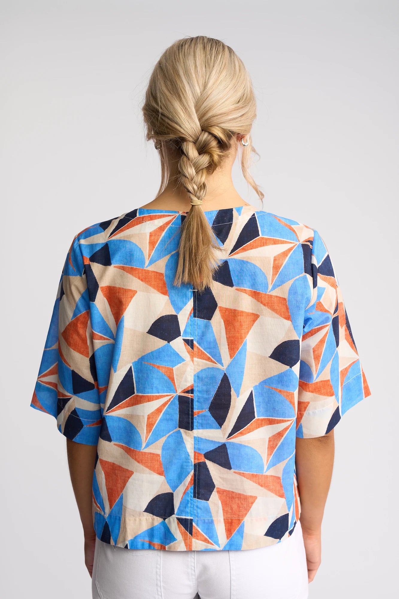 A blonde-haired person with a braid wears the Vassalli Athena Top - Row, featuring a vibrant geometric print in blue, orange, and beige hues. The cotton blouse has a boxy fit and is showcased as they face away from the camera against a plain background.