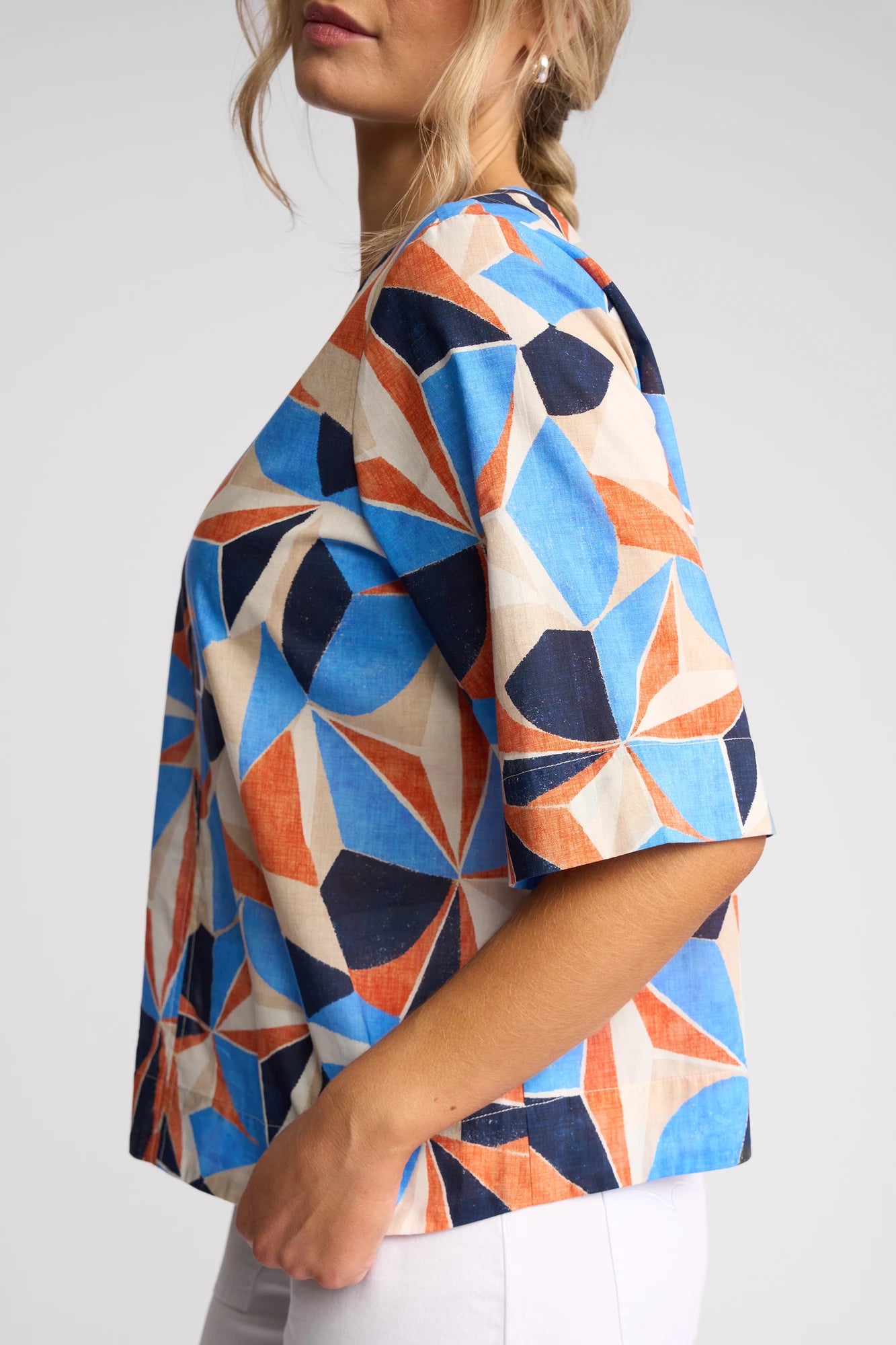 A woman stands sideways against a plain background with her hair in a loose braid, wearing the Vassalli Athena Top - Row. The cotton blouse showcases a colorful geometric print with blue, orange, and beige shapes, and its boxy fit is complemented by light-colored pants.