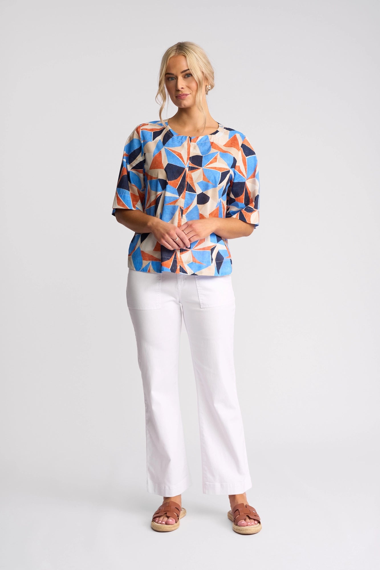 A woman stands against a plain background, wearing the Vassalli Athena Top - Row, which features a geometric print in blue, orange, and beige. The cotton top has a boxy fit that pairs effortlessly with white pants and brown sandals. She has blonde hair styled in a loose updo and her hands are clasped in front.
