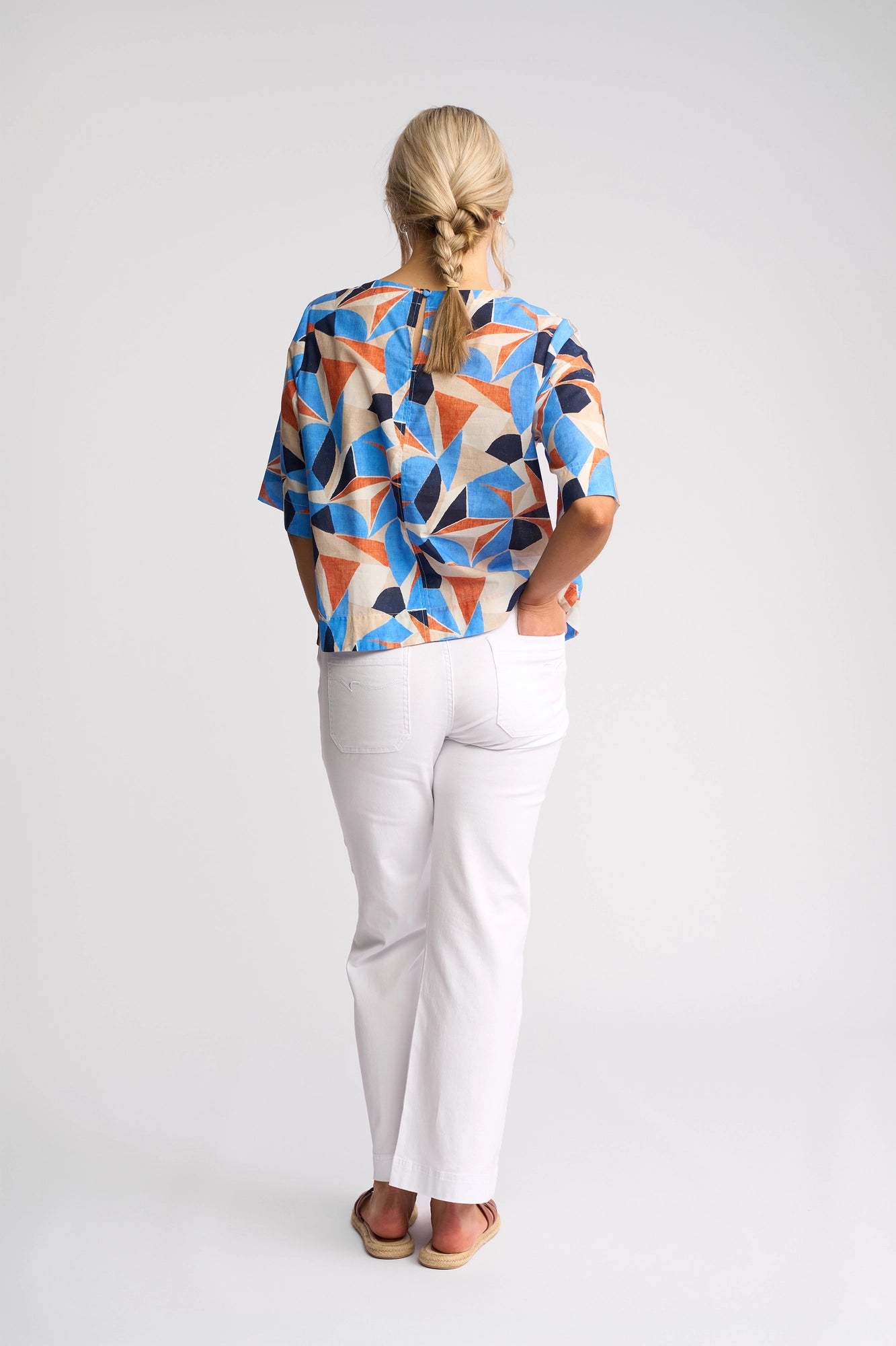 A woman with blonde hair in a braid stands facing away, wearing the Vassalli Athena Top - Row, featuring a vibrant geometric print and a boxy cotton fit, paired with white pants and sandals, her hands casually tucked in her pockets against a plain white background.