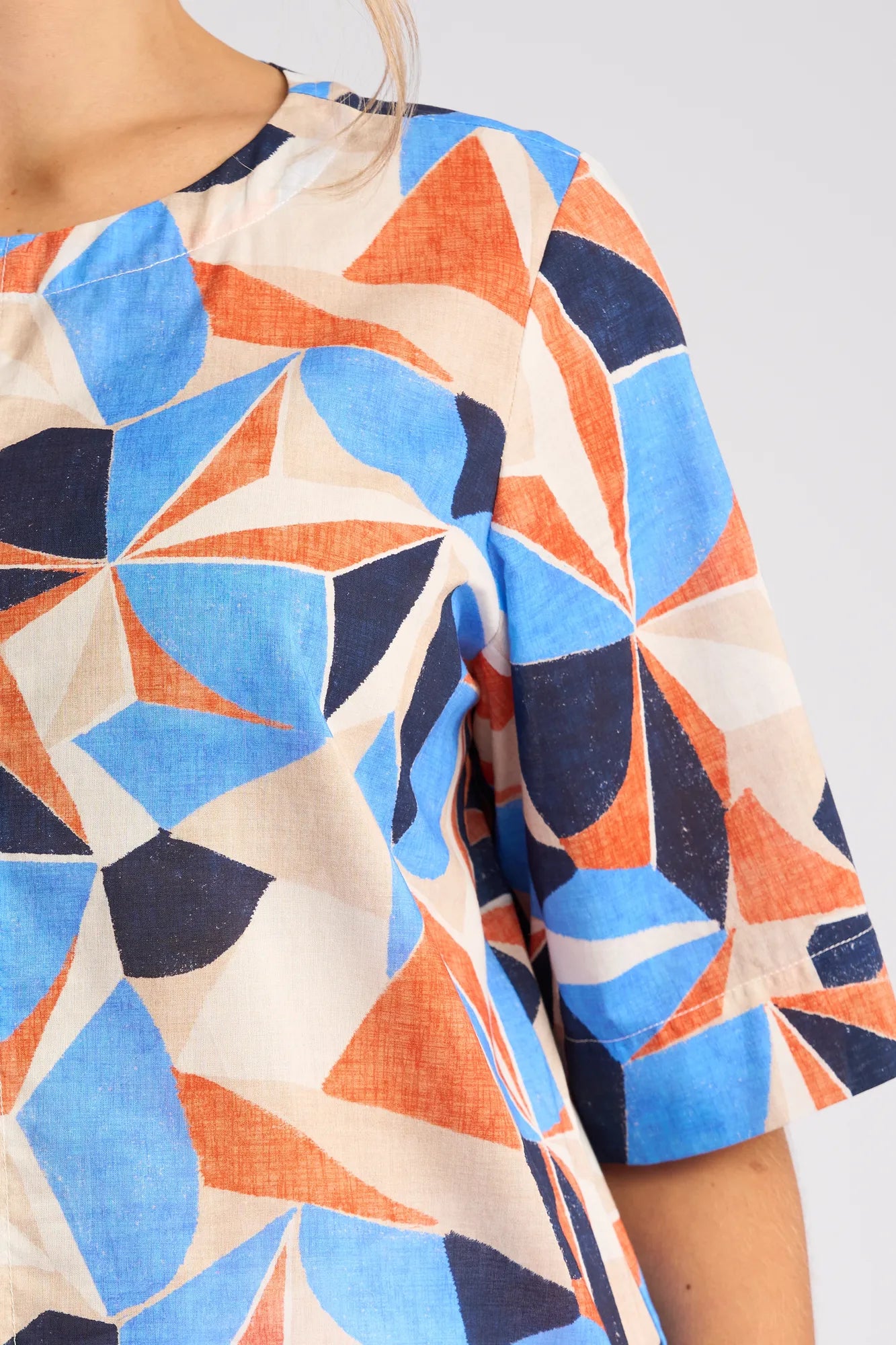 A close-up of a person wearing the Vassalli Athena Top - Row, featuring a striking geometric print in blue, orange, navy, and beige. This cotton shirt offers a boxy fit with short sleeves and a round neckline. Only the upper torso and part of the arm are visible.