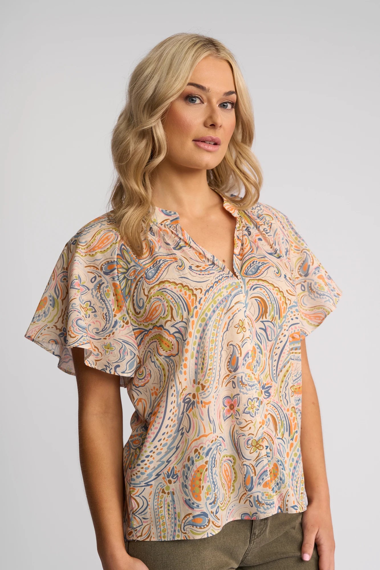 A woman with blonde hair wears the Vassalli Nyla Top - Aurora, a short-sleeved cotton blouse featuring a pastel paisley print. She stands against a plain light gray background, gazing slightly to the side with a relaxed expression.