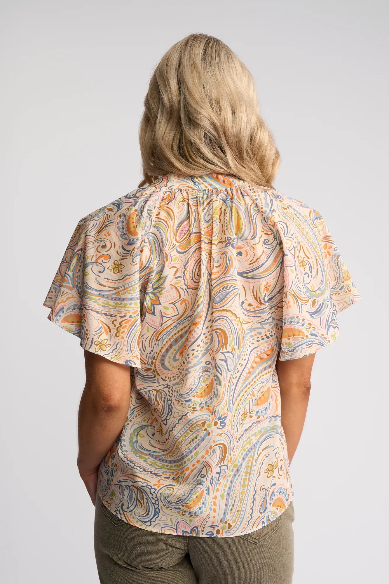 A person with long blonde hair, seen from behind, wears the Vassalli Nyla Top - Aurora, a light cotton blouse featuring a colorful paisley print, paired with olive green pants against a plain, light background.