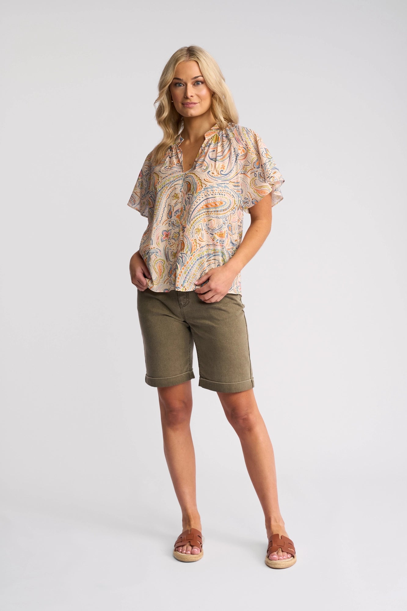 A woman with wavy blonde hair stands against a plain background, wearing the Vassalli Nyla Top – Aurora, olive knee-length shorts, and sandals. She has one hand in her pocket and is looking at the camera.