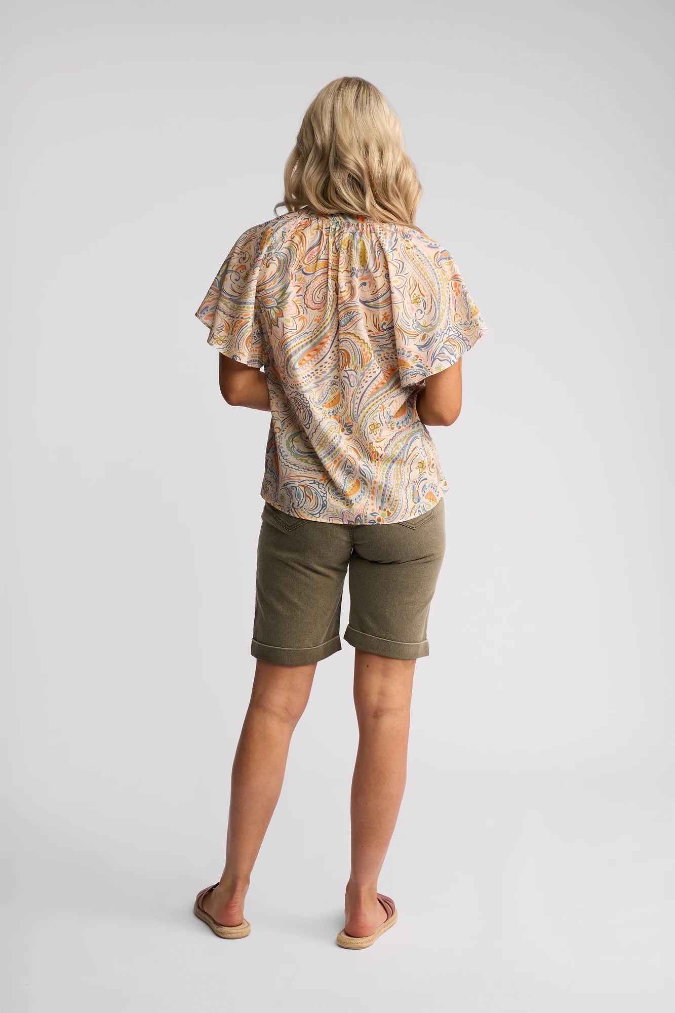A woman with blonde hair stands facing away, wearing the Vassalli Nyla Top - Aurora, a colorful paisley print cotton blouse with flutter sleeves, paired with olive green knee-length shorts and sandals, against a plain light background.