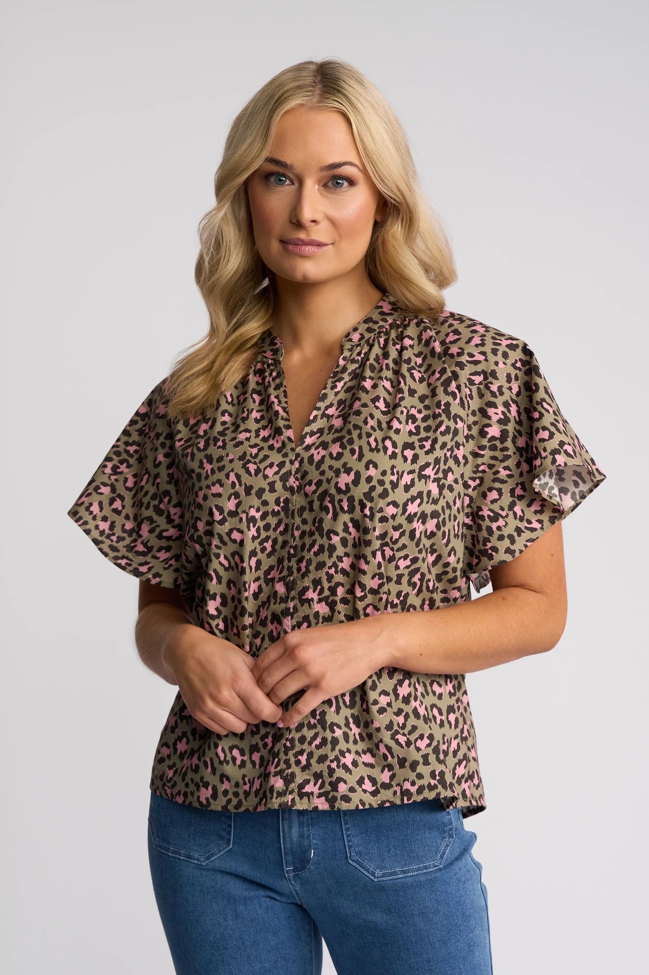 A woman with long blonde hair wears the Vassalli Nyla Top - Cub, a short-sleeved, v-neck leopard print top with pink spots, paired with blue jeans and standing against a plain light background.