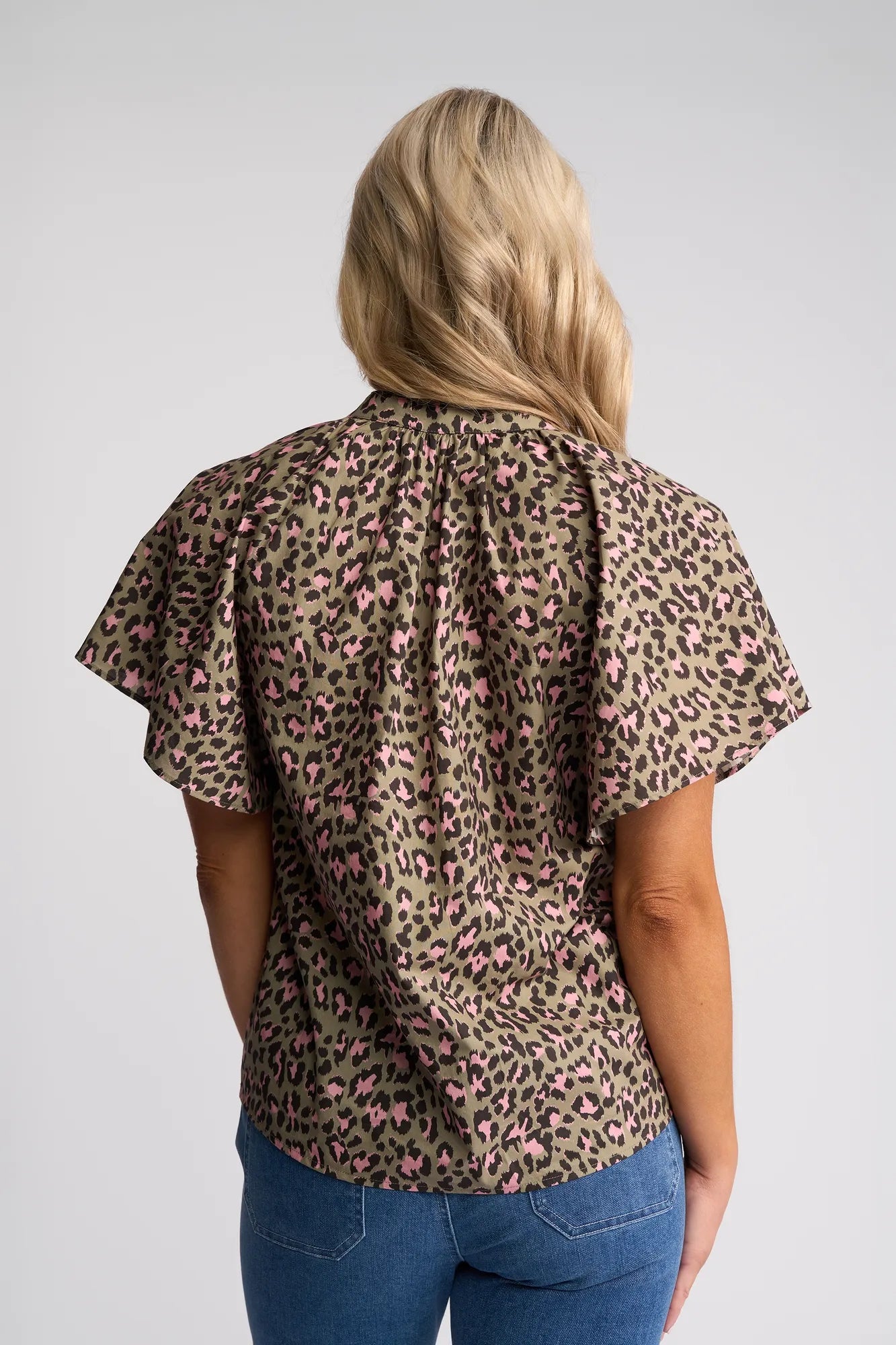 A woman with long blonde hair stands facing away from the camera against a plain background, wearing the Vassalli Nyla Top - Cub—a loose, short-sleeved leopard print top in brown, black, and pink with subtle flutter sleeves—paired with blue jeans.