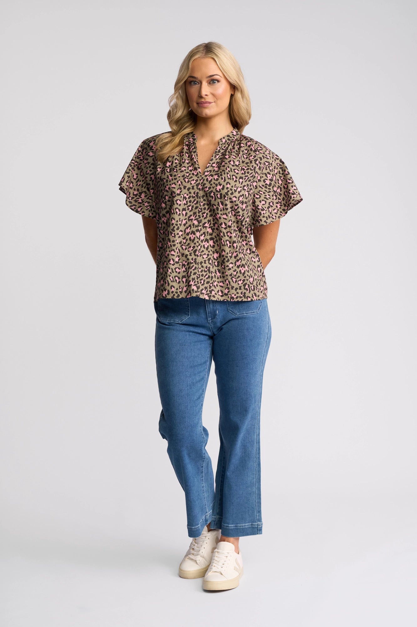 A woman stands in a studio wearing the Vassalli Nyla Top - Cub, paired with blue jeans and white sneakers. She has long blonde hair, a neutral expression, and her hands are loosely resting behind her back.