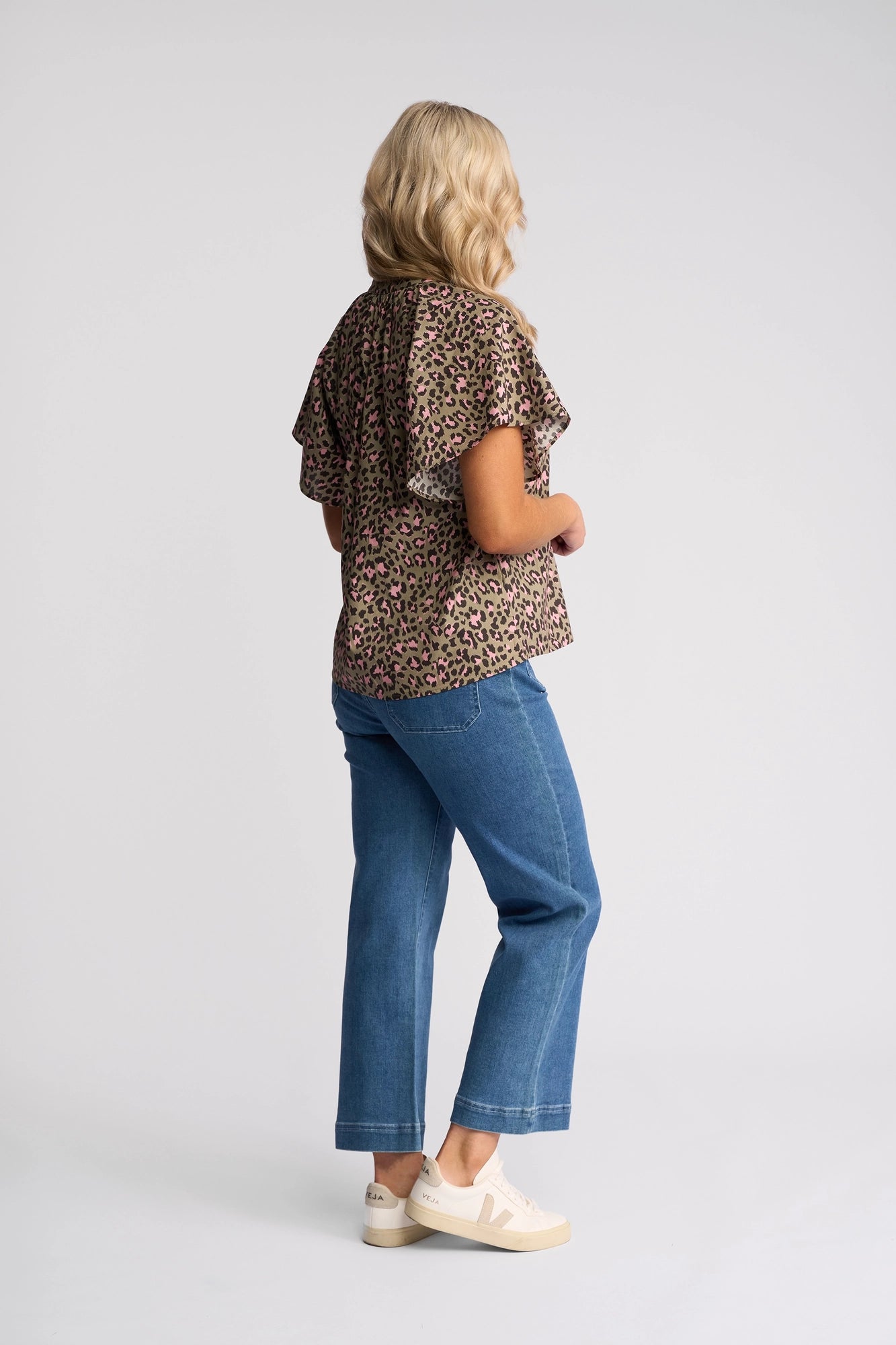 A woman with long blonde hair, wearing the Vassalli Nyla Top - Cub featuring a leopard print and flutter sleeves, paired with blue jeans and white sneakers, stands facing away in a studio with a plain light background.