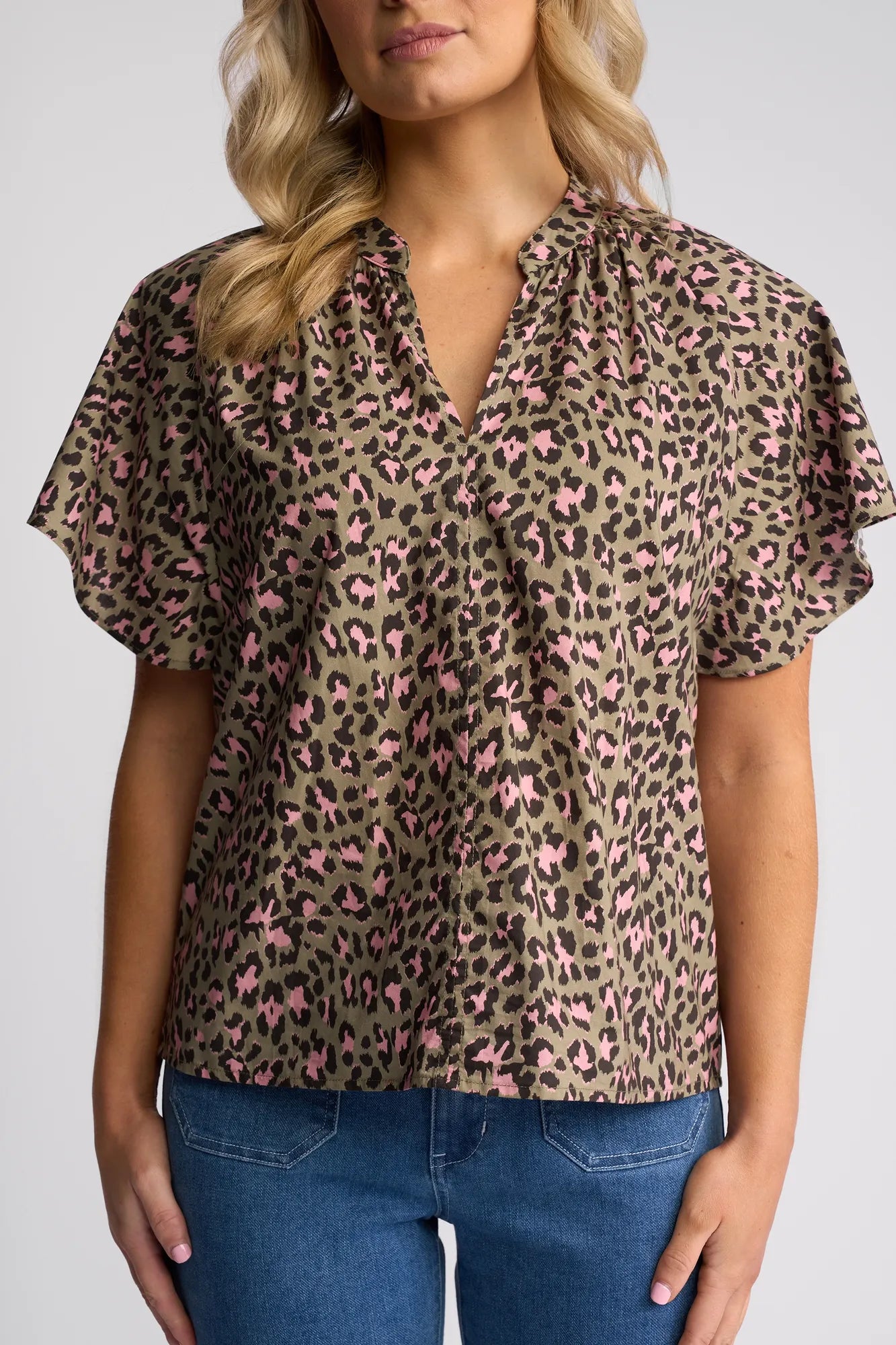 A woman wears the Vassalli Nyla Top - Cub, a short-sleeve, V-neck cotton blouse featuring a leopard print with pink and black spots on olive green fabric. She has wavy blonde hair and pairs the top with blue jeans. The photo is cropped at the thighs.