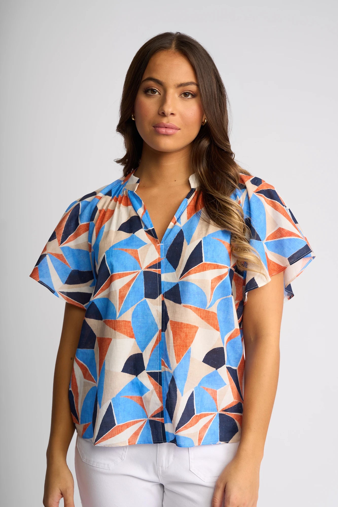A woman wearing the Vassalli Nyla Top - Row, a short-sleeved, v-neck cotton blouse featuring an abstract geometric pattern in blue, orange, and white, stands against a plain light background. She has long wavy hair and a neutral expression.