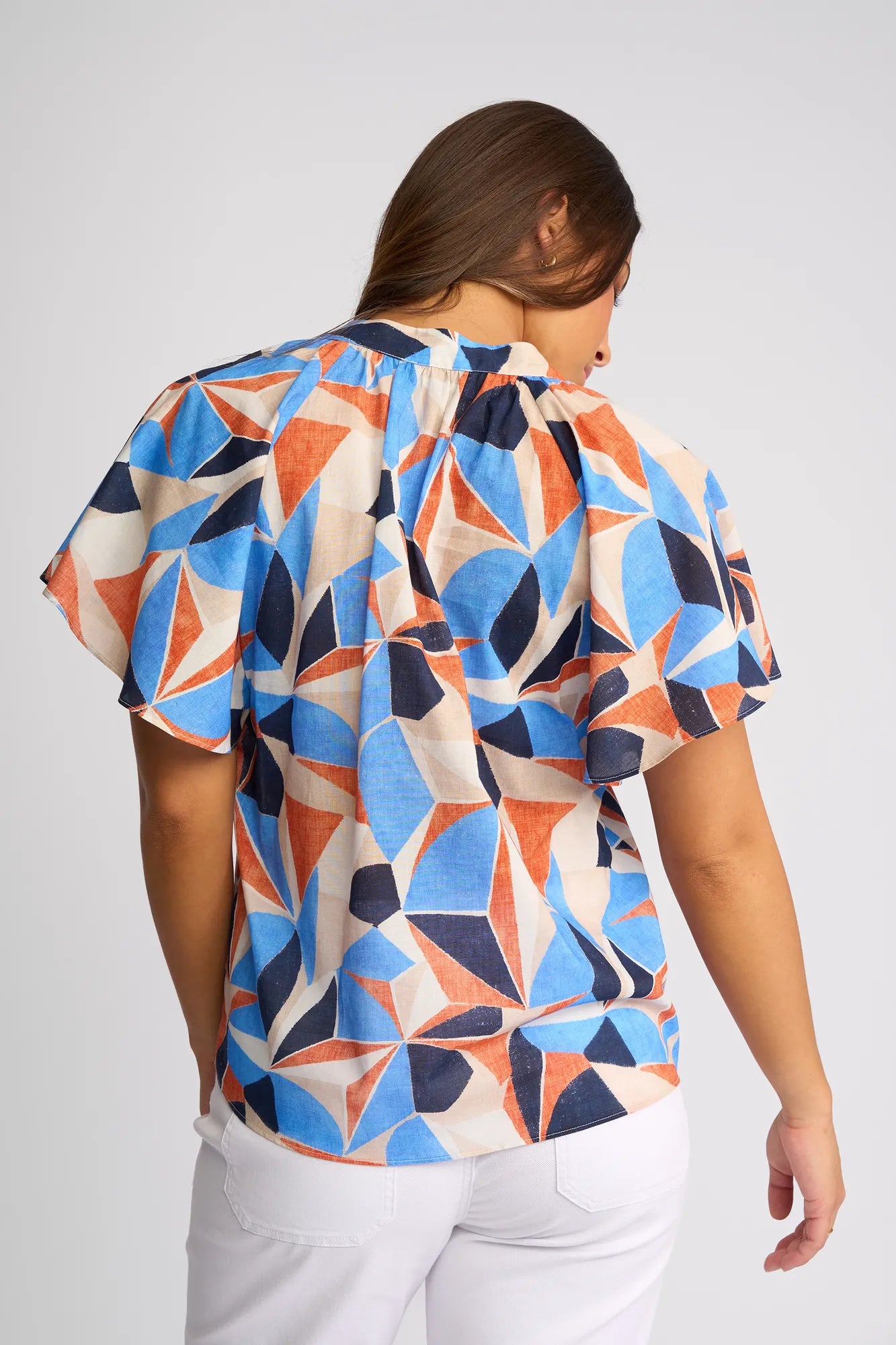 A woman with brown hair, seen from behind, wears the Vassalli Nyla Top - Row—a short-sleeve cotton blouse featuring a vibrant blue, orange, and beige geometric print. She pairs it with white pants against a plain light background.