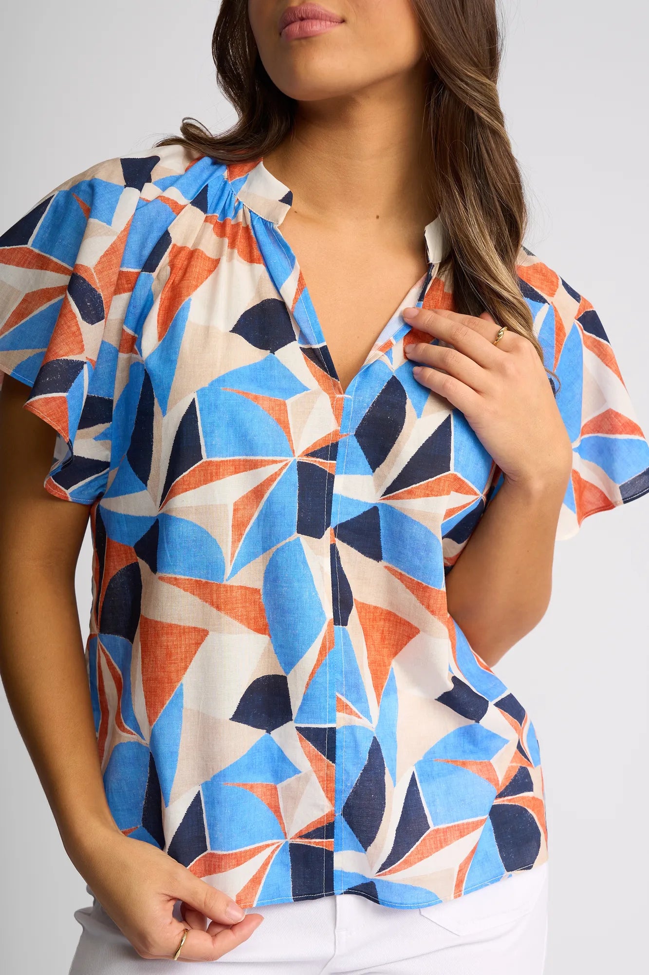 A woman wearing the Vassalli Nyla Top - Row, a colorful geometric print cotton blouse with flutter sleeves in blue, orange, and cream shapes, paired with white pants. She is touching the collar with one hand while standing against a plain background.