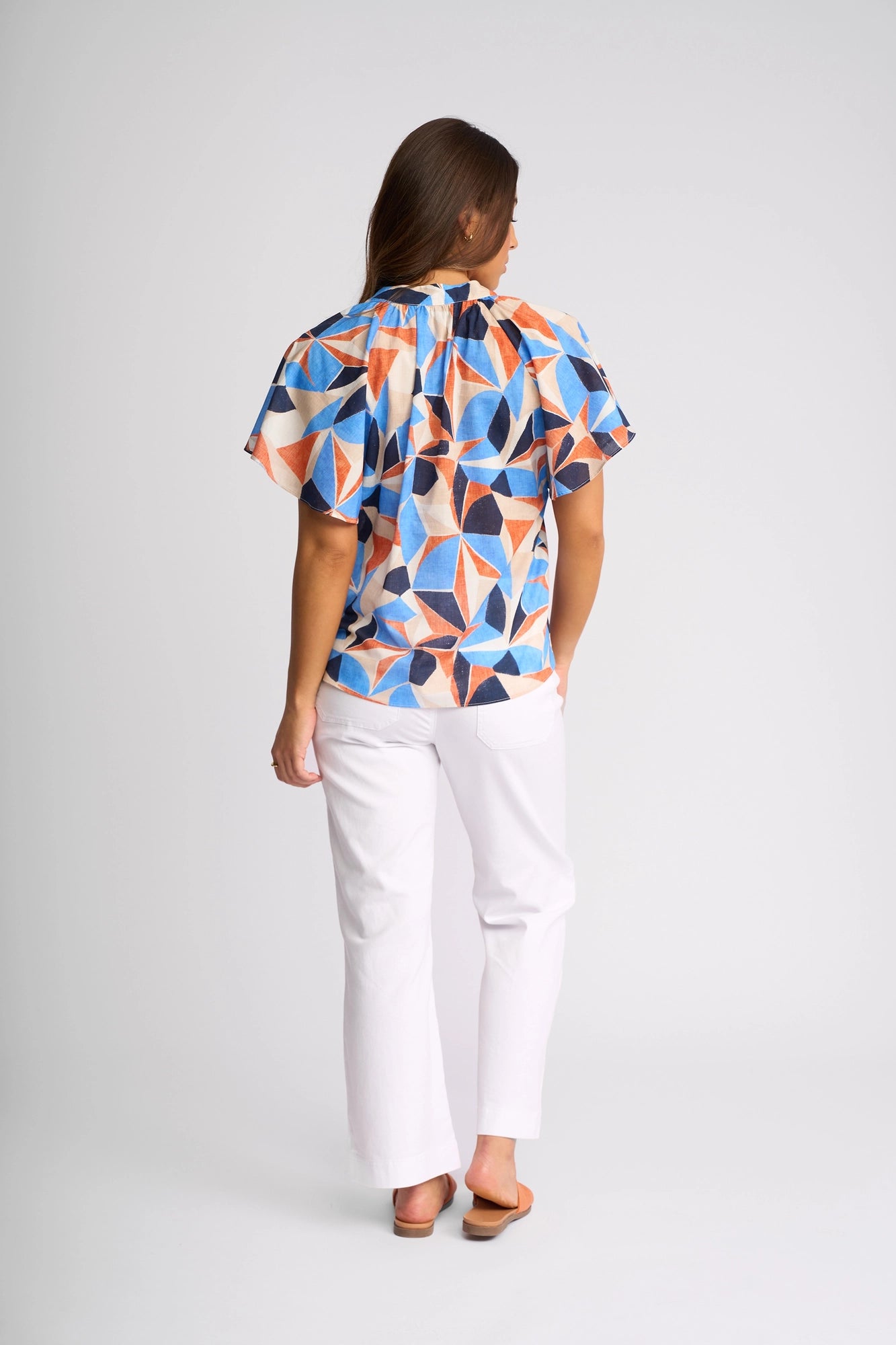 A person stands facing away, wearing the Vassalli Nyla Top - Row, a vibrant geometric print cotton blouse with flutter sleeves, paired with white pants and brown sandals against a plain, light gray background.