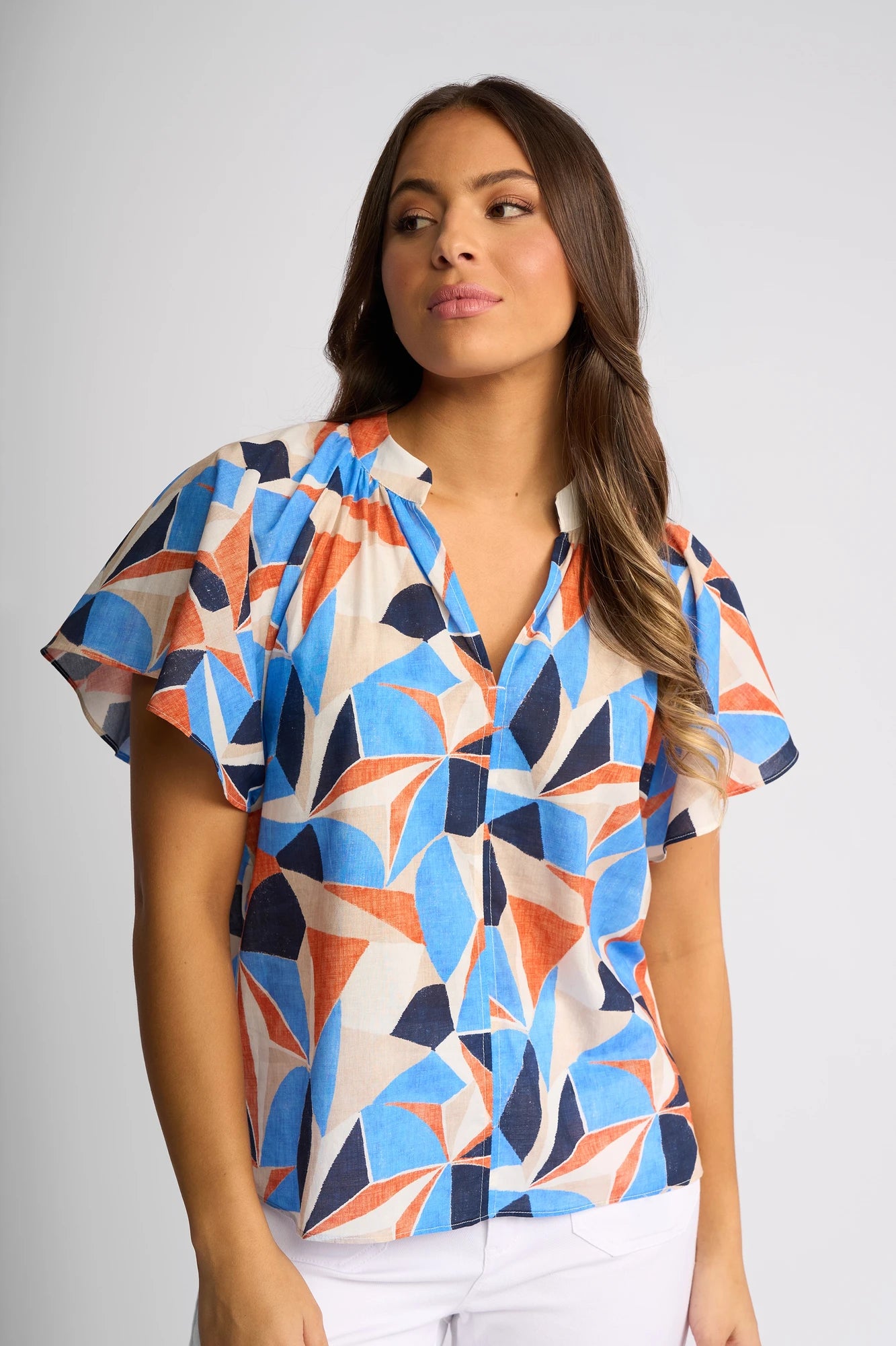 A woman wearing the Vassalli Nyla Top - Row, featuring a blue, orange, and cream geometric print cotton blouse, looks to the side while standing against a plain light background.