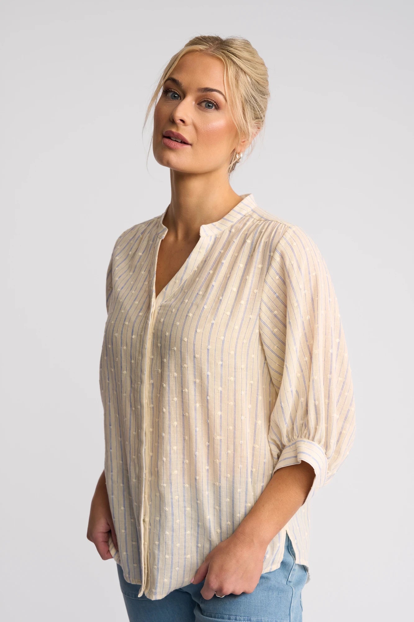 A woman stands against a plain white background, gazing slightly upward with a relaxed expression, wearing the Vassalli Folk Shirt - Blue Stripe and blue jeans. The light, long-sleeve shirt is crafted from textured cotton, bringing effortless style to her laid-back look.