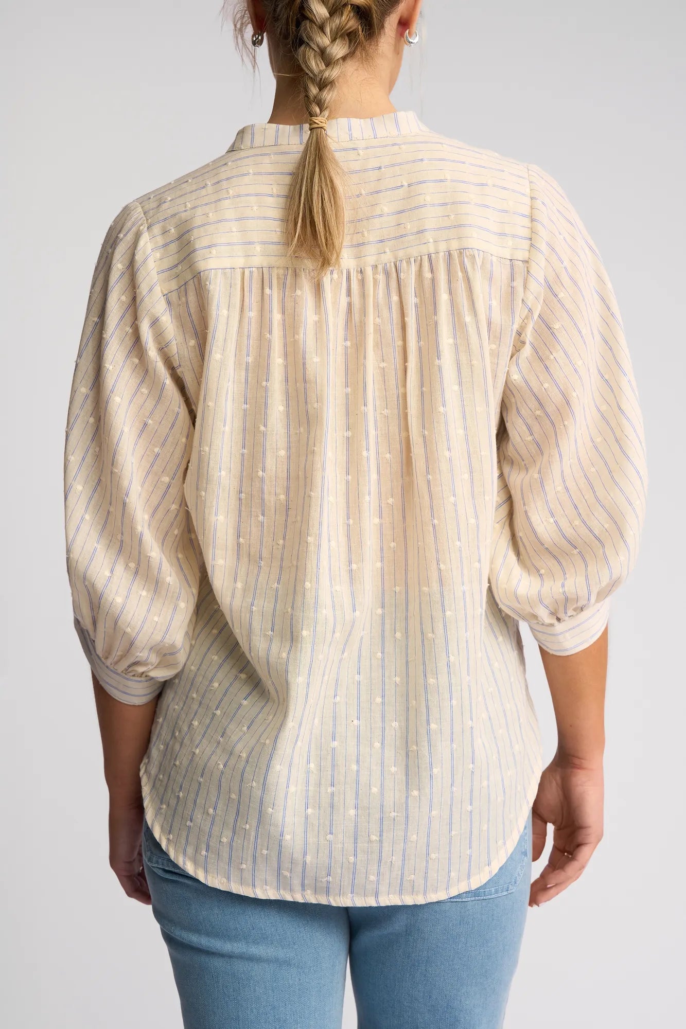 A person with a blonde braid is seen from the back wearing the Vassalli Folk Shirt in Blue Stripe, featuring light, semi-sheer fabric with puffed sleeves, paired with light blue jeans against a plain background.