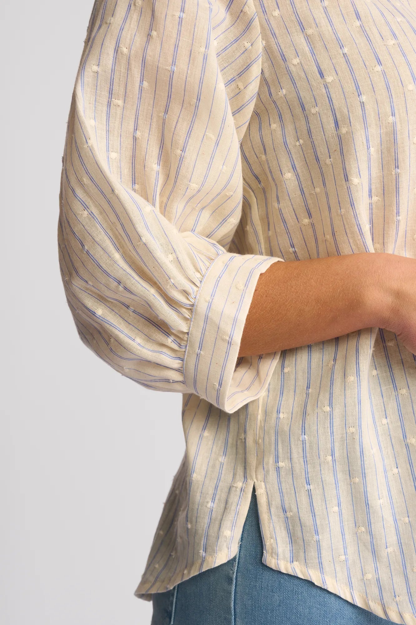A person is wearing the Vassalli Folk Shirt - Blue Stripe—a cream blouse adorned with blue vertical stripes and textured cotton dots. The puff-sleeved top, crafted from textured cotton by Vassalli, is styled with light blue jeans. Only the torso and arm are visible.