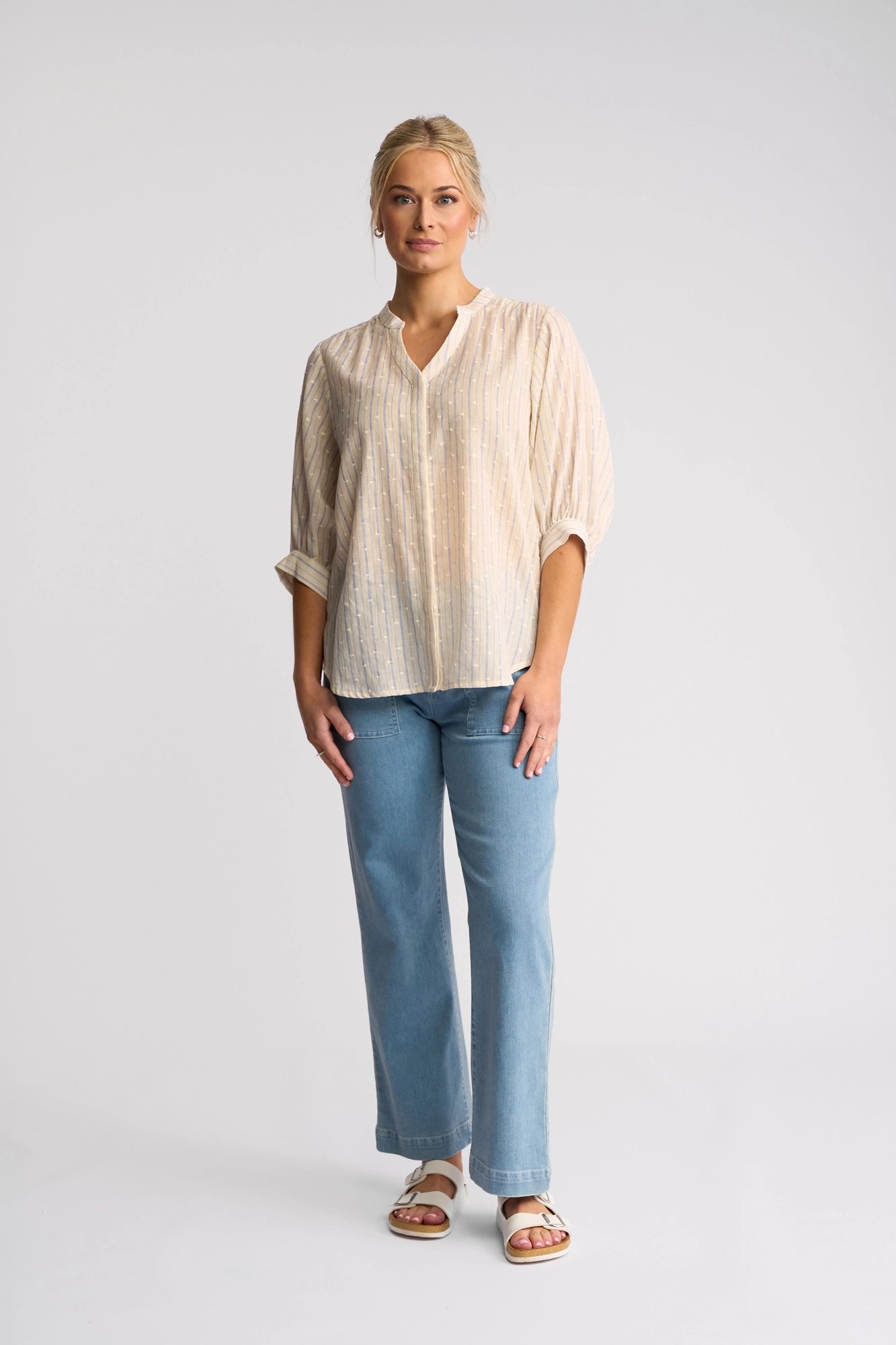 A woman stands against a plain background, wearing a Vassalli Folk Shirt in Blue Stripe with long sleeves, paired with light blue jeans and white sandals. She has blonde hair pulled back and a relaxed, neutral expression.