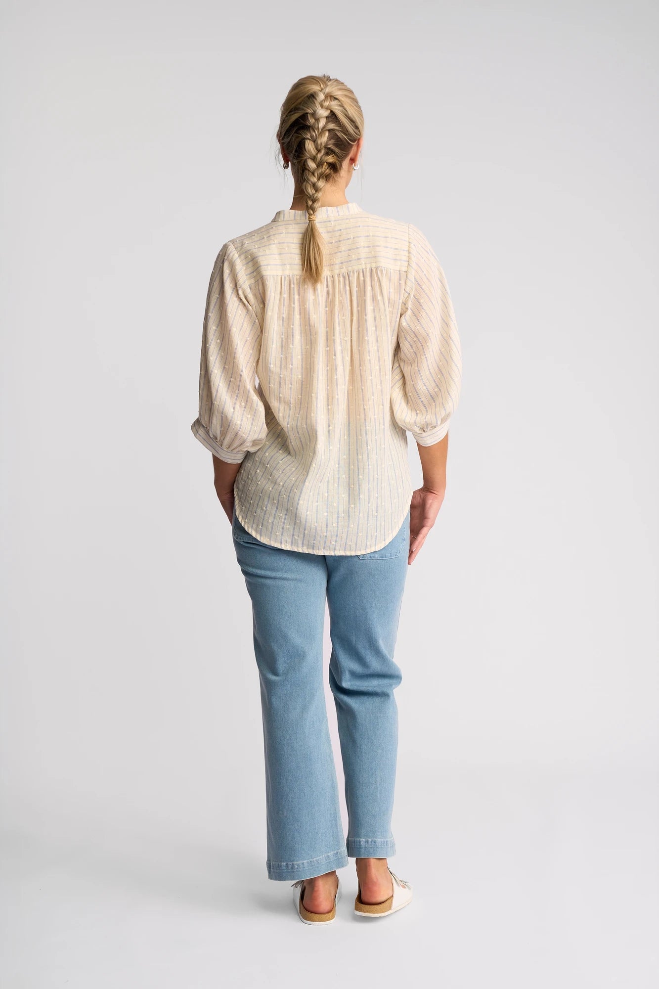 A person with a blonde braid, wearing a Vassalli Folk Shirt in Blue Stripe, blue jeans, and beige shoes, stands facing away against a plain white background.