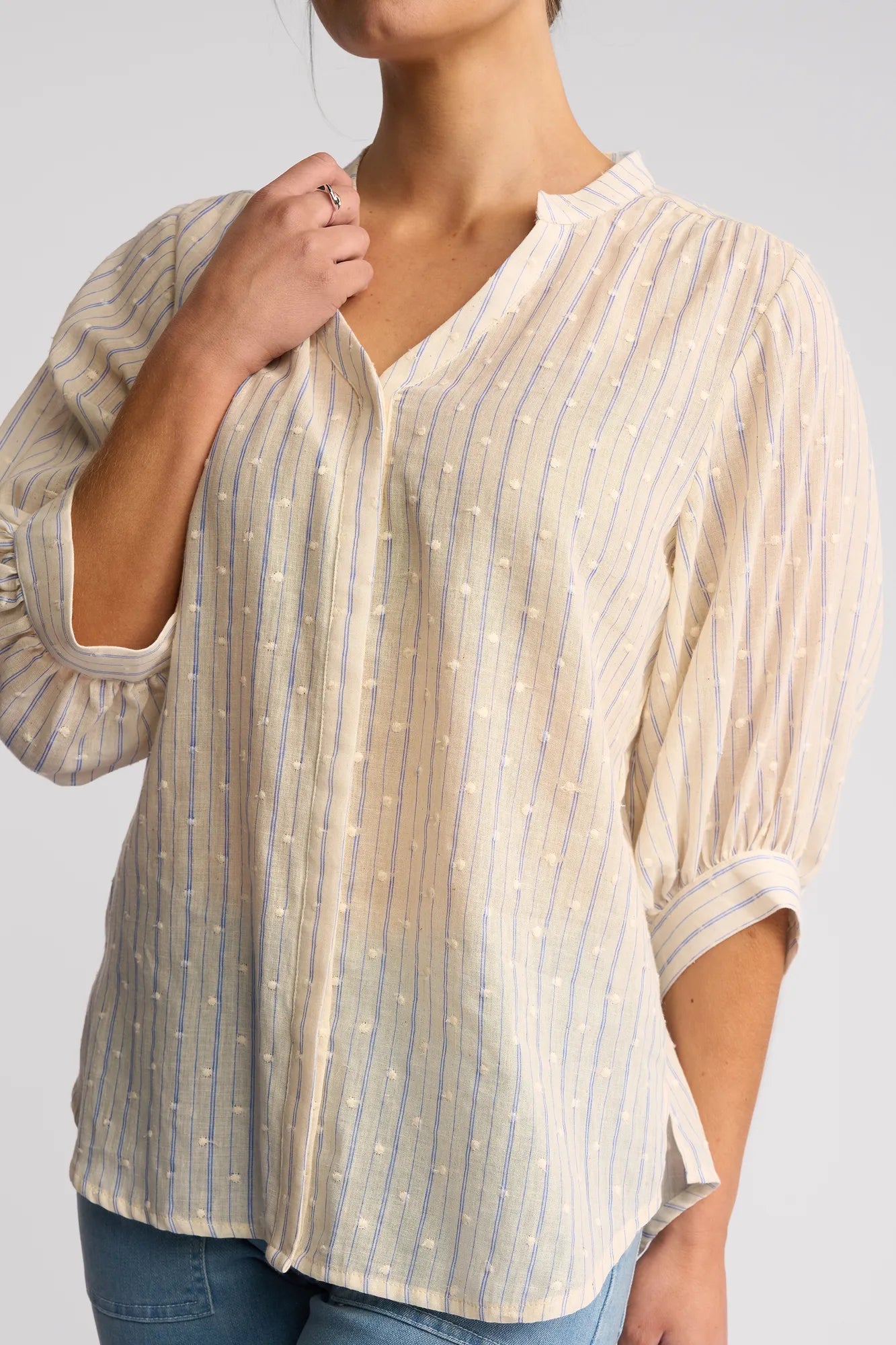 A woman wears the Vassalli Folk Shirt - Blue Stripe, featuring long sleeves with vertical blue stripes and subtle dot patterns, paired with blue jeans. Her left hand rests on her chin.
