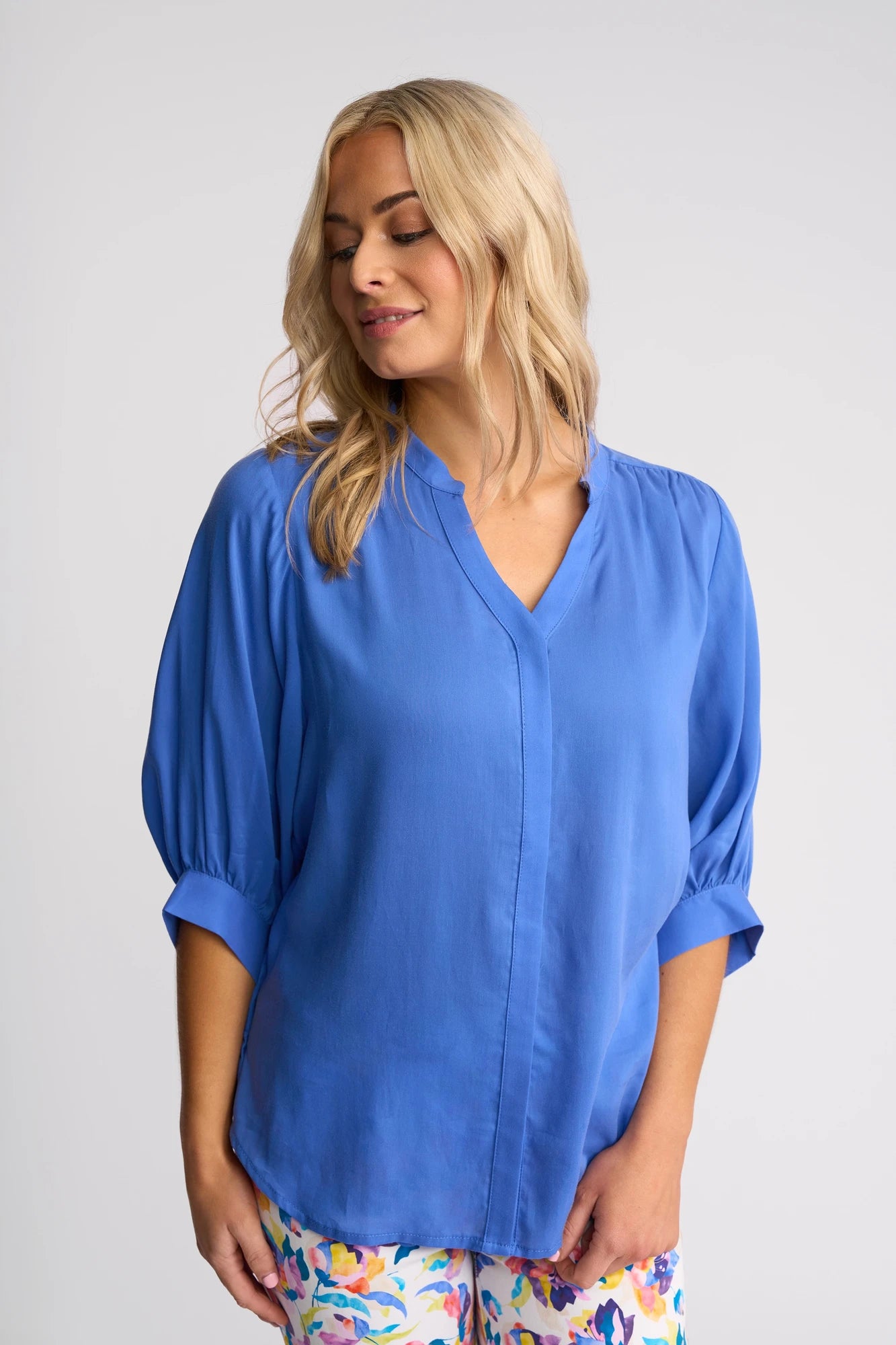 A blonde-haired woman stands against a plain light background, wearing the Vassalli Folk Shirt in French Blue—a loose-fitting Tencel top with three-quarter sleeves—paired with bright, colorful floral pants.
