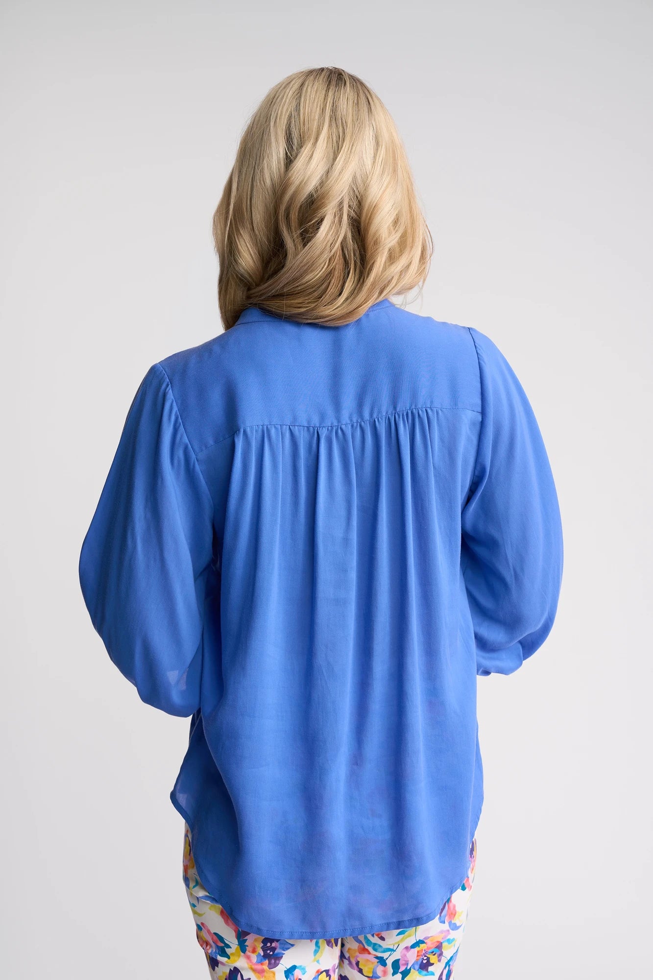 A person with wavy blonde hair, dressed in the Vassalli Folk Shirt - French Blue and patterned pants, stands with their back to the camera against a plain light background.