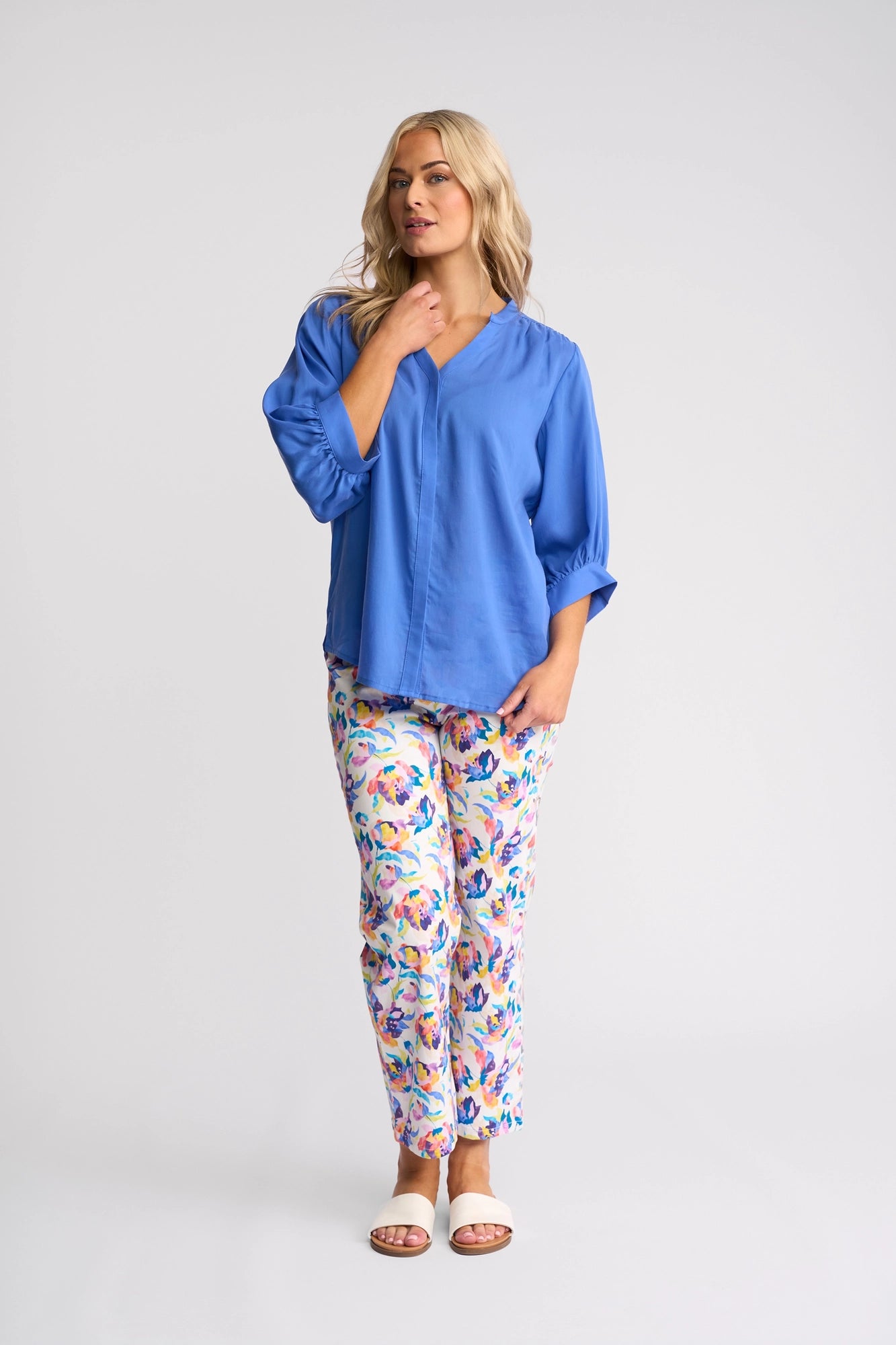 A woman with long blonde hair wears the Vassalli Folk Shirt in French Blue, pairing it with colorful floral pants and white sandals. She stands against a plain light background, looking to the side with one hand near her chin.