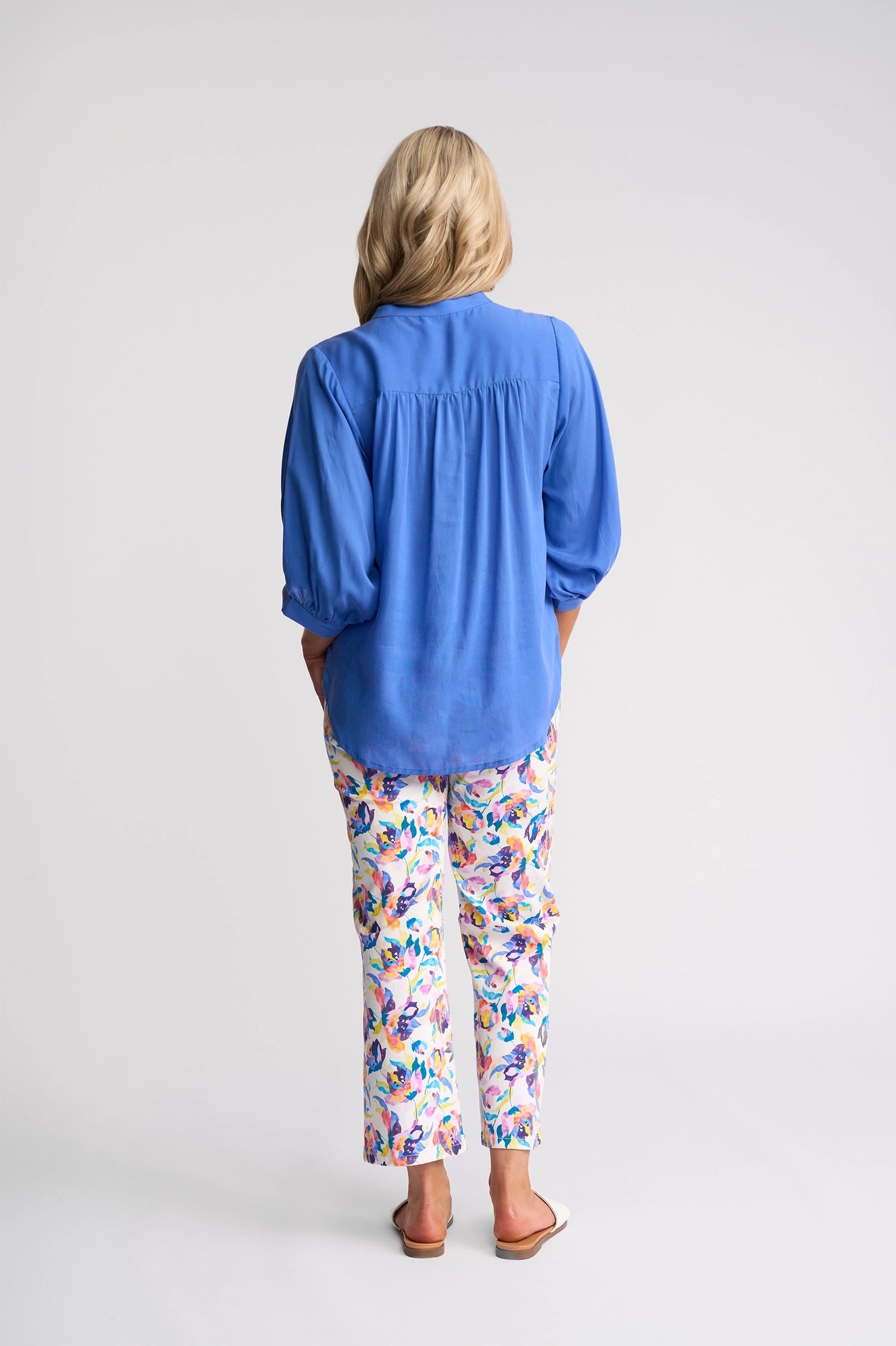A person with long blonde hair faces away, wearing the Vassalli Folk Shirt in French Blue, paired with colorful floral pants and white sandals, standing against a plain white background.