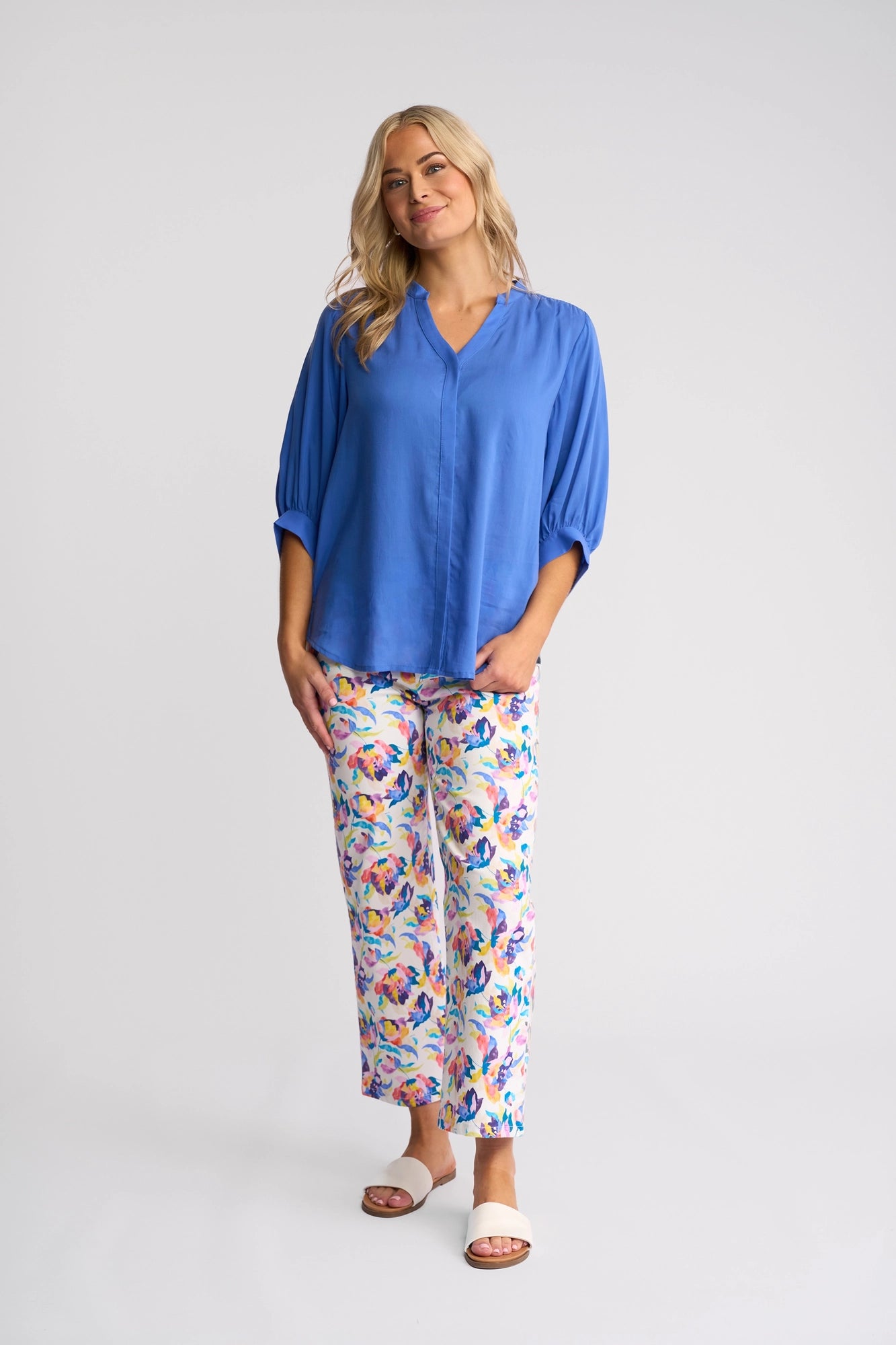 A woman stands and smiles, wearing the Vassalli Folk Shirt in French Blue, paired with colorful floral print pants and white sandals against a plain light background.