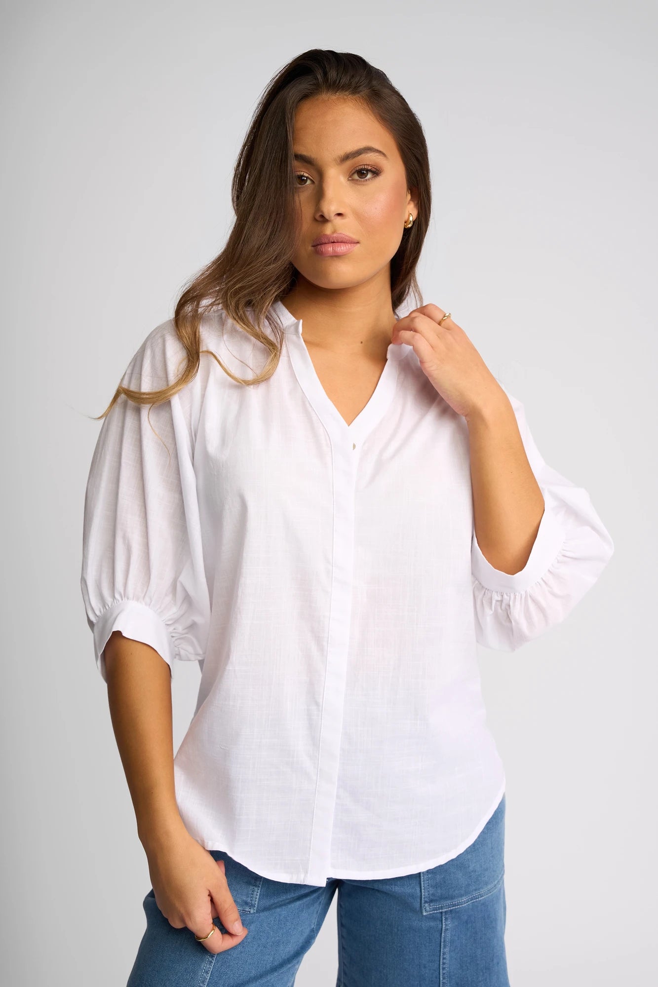 A woman with long brown hair, dressed in the Vassalli Folk Shirt - White—a lightweight cotton top featuring three-quarter sleeves and a relaxed silhouette—paired with blue jeans, stands against a plain light gray background, looking confidently at the camera.
