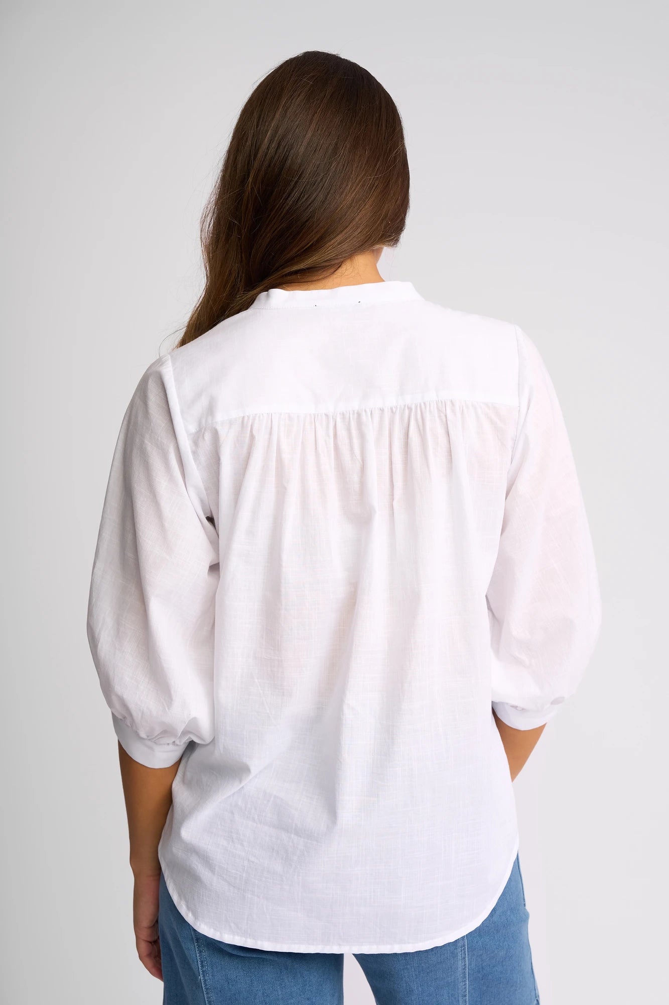 A person with long brown hair is shown from behind, wearing the Vassalli Folk Shirt in white—a relaxed silhouette featuring three-quarter sleeves—paired with light blue jeans, standing against a plain background.
